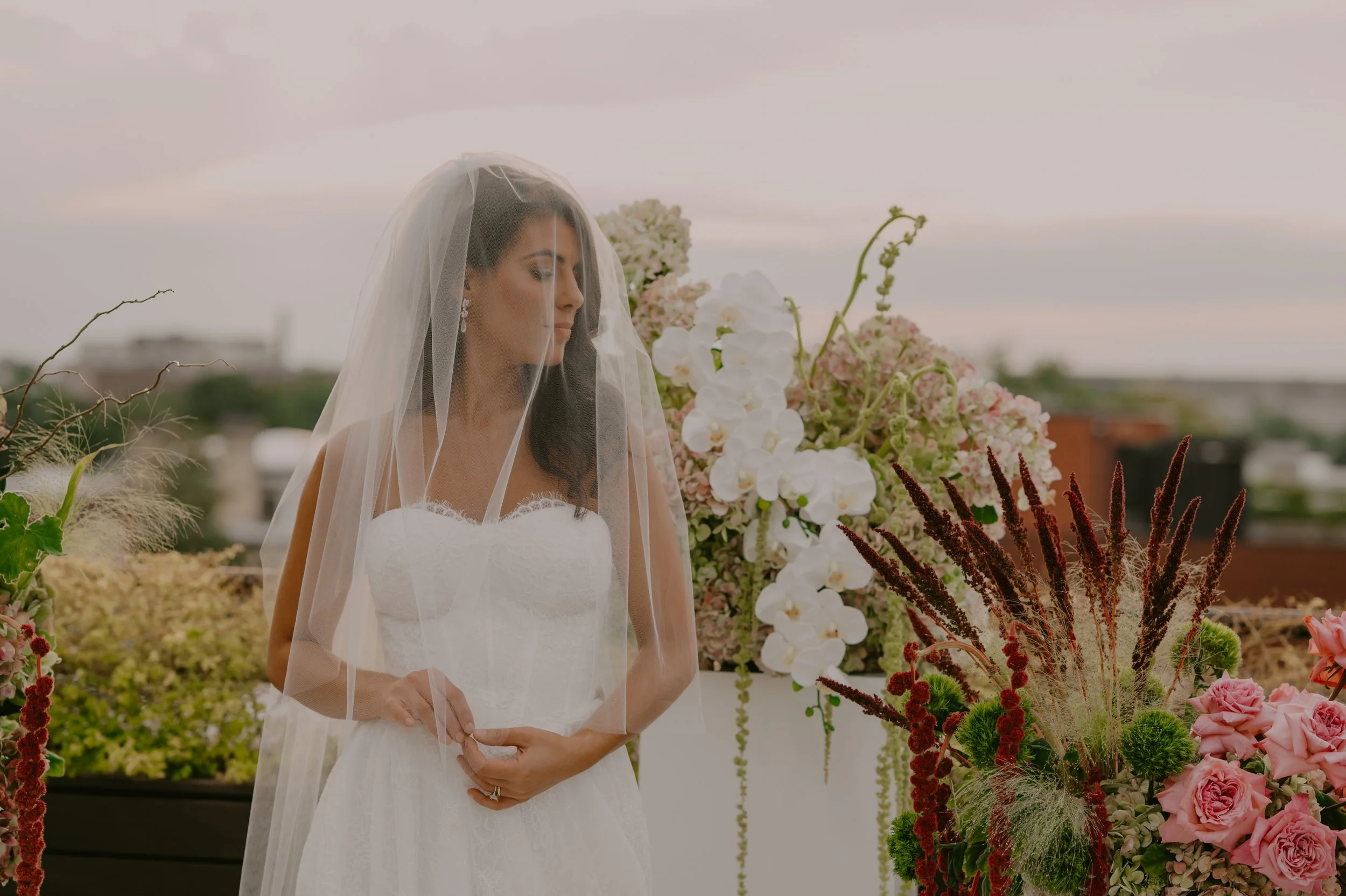 Bride in a white wedding dress and veil standing near colorful floral arrangements on a rooftop with cityscape in the background.