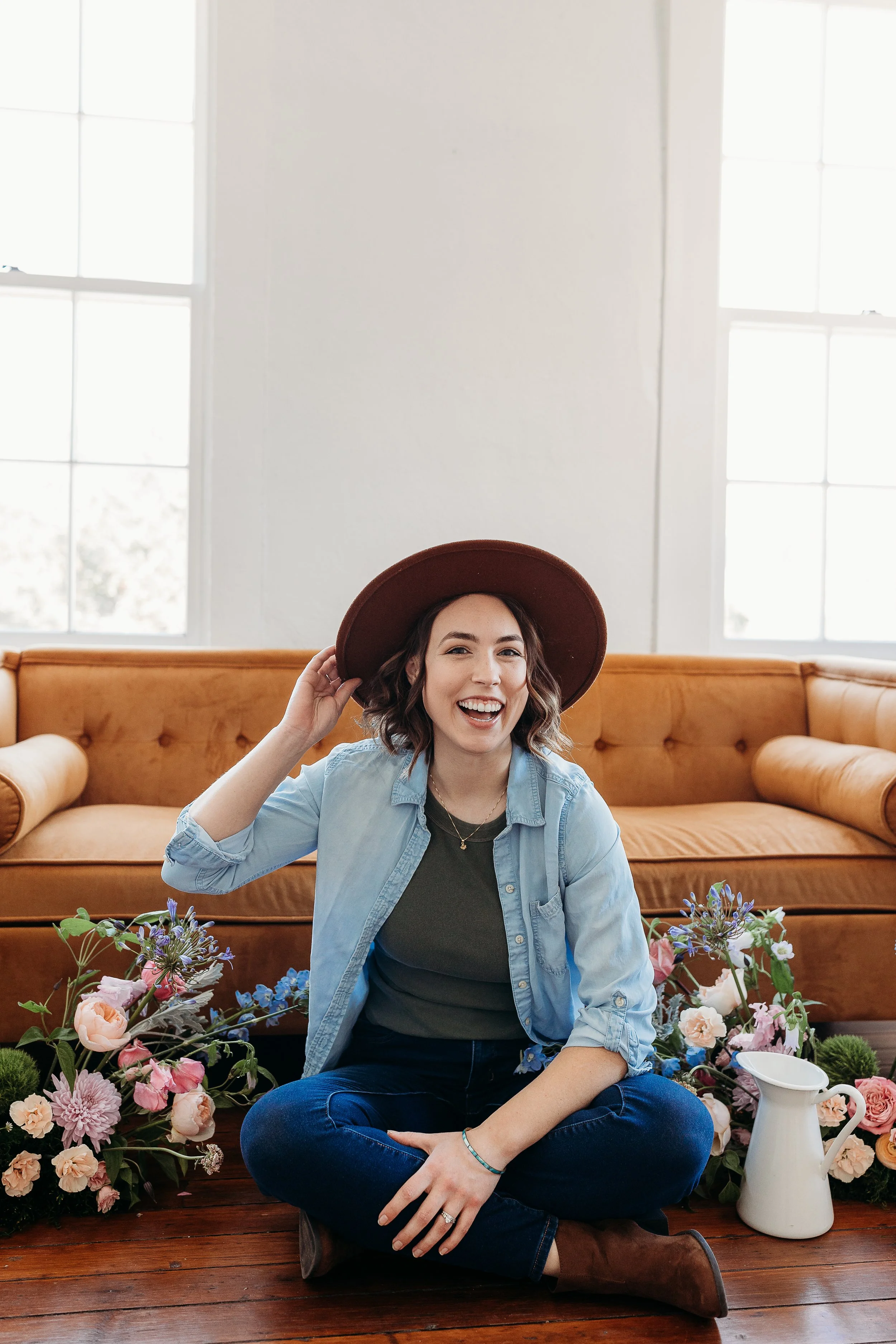 A young woman sitting cross-legged on a wooden floor, smiling, wearing a blue denim shirt and a dark green top, with a wide-brimmed brown hat. She is surrounded by pink, purple, and white flowers, and there is a white pitcher nearby. She is indoors, with large windows and a peach-colored sofa in the background.
