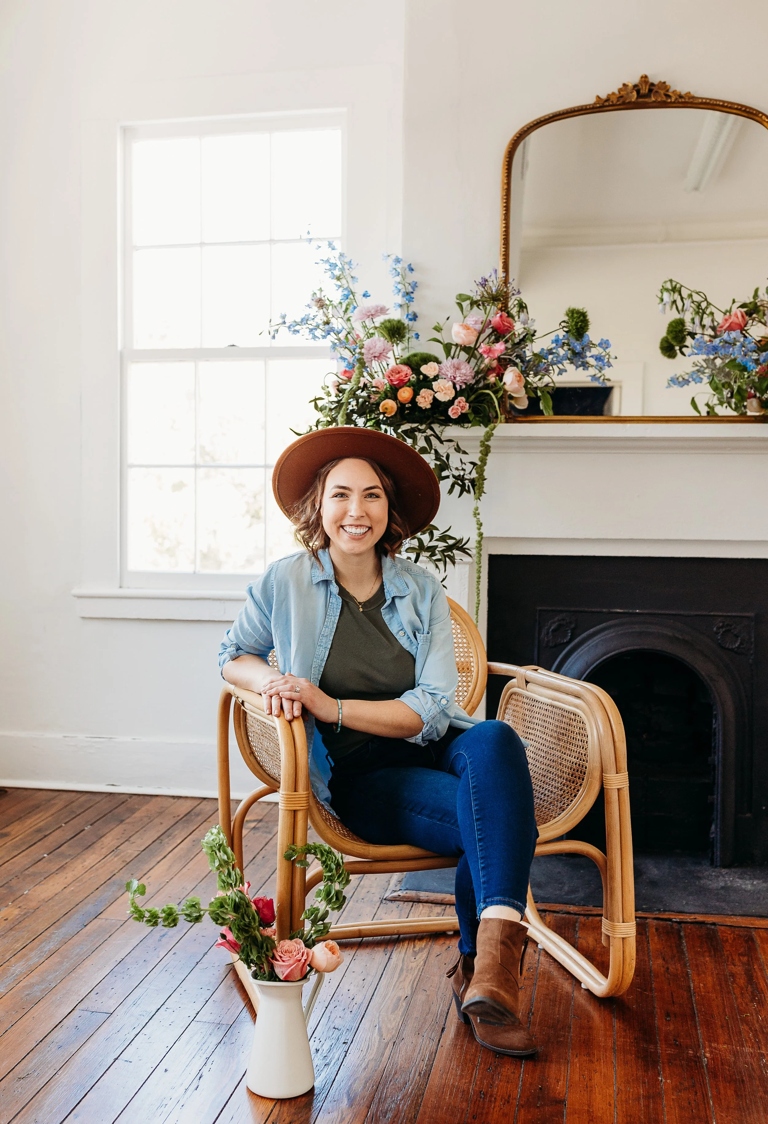A woman sitting in a wooden chair in a room with a fireplace and a large window, smiling while wearing a wide-brimmed hat, denim shirt, and dark jeans, surrounded by colorful flowers.