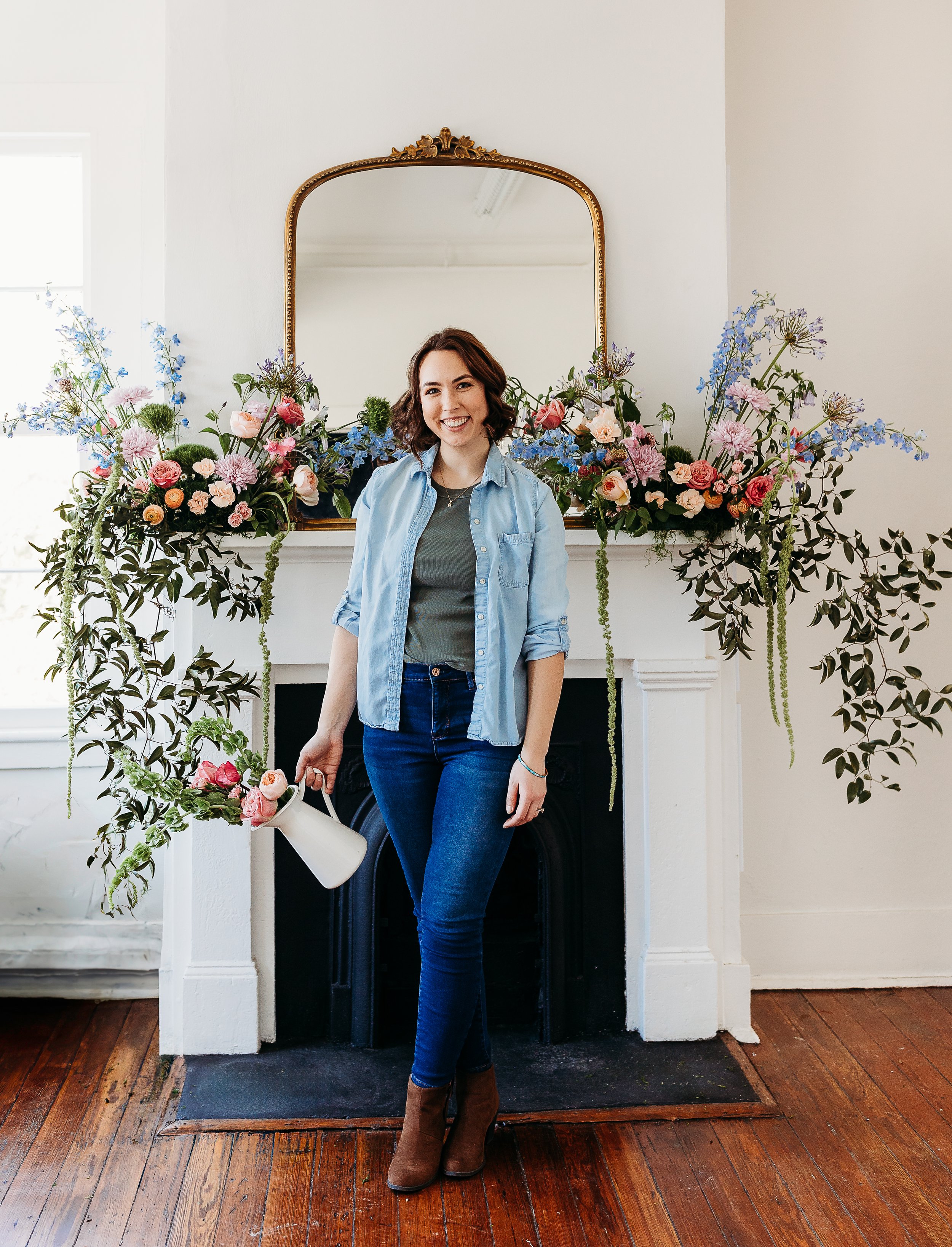 A woman with shoulder-length curly brown hair, wearing a light blue denim shirt over a green top, blue jeans, and brown ankle boots, stands in front of a fireplace decorated with an elaborate arrangement of pink, purple, and blue flowers, with a gold-framed mirror above it. She is smiling and holding a white pitcher with pink flowers in it.