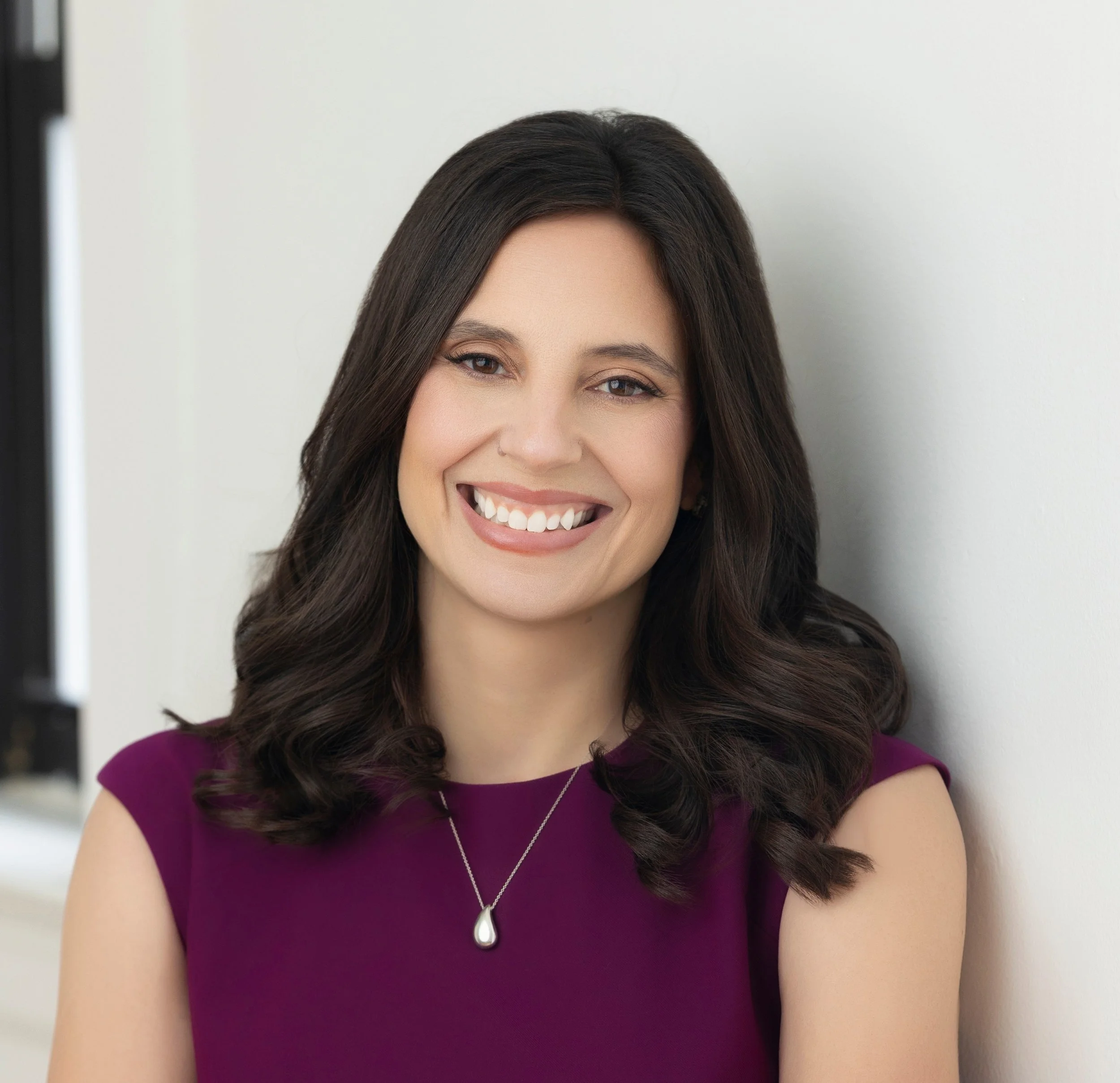 Portrait of Nicole Shannon wearing a purple dress standing against a white wall.