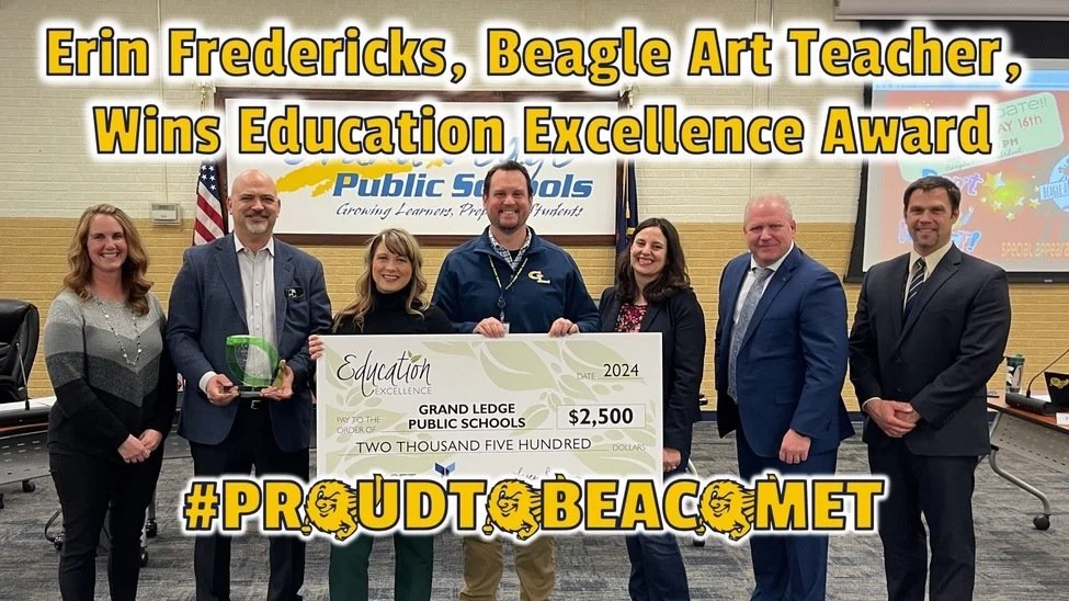 Group of seven people standing inside a room, holding a large check for $2,500, with a screen in the background displaying the same information, celebrating Erin Fredericks, a Beagle Art Teacher, winning the Education Excellence Award at Grand Ledge Public Schools.