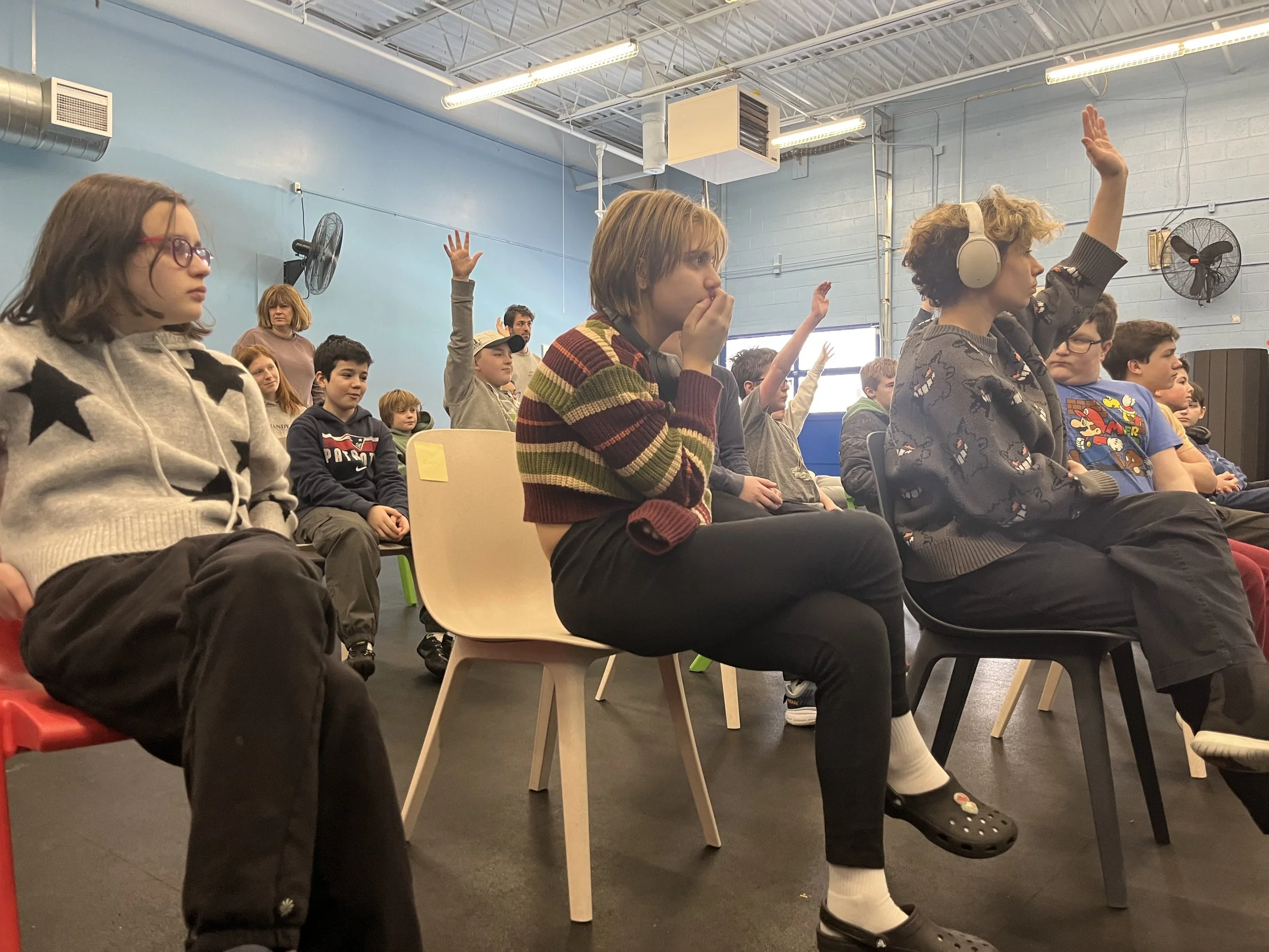 Children seated in a classroom, raising hands, listening to a lesson or presentation.