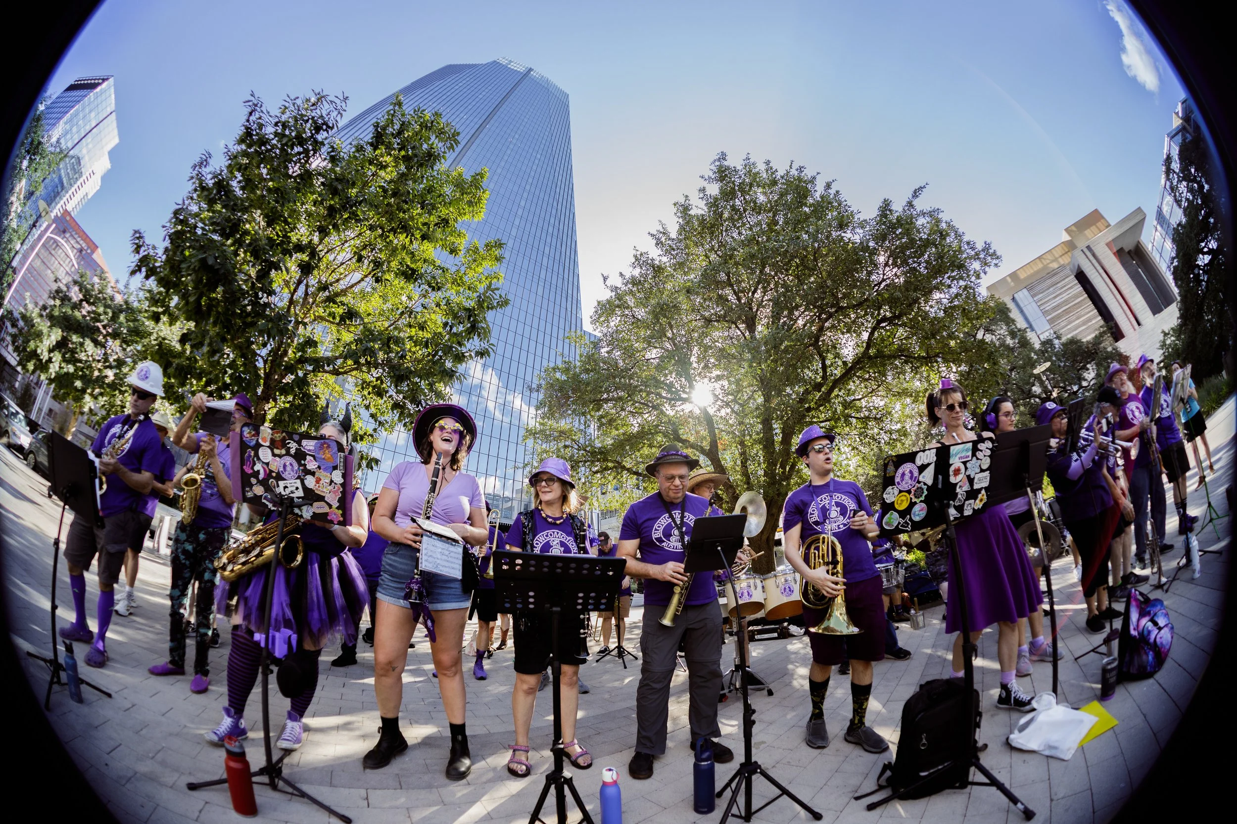 Members of Austin brass ensemble Blowcomotion perform in purple shirts at Republic Square, framed by trees and downtown towers during HAAM Day 2025