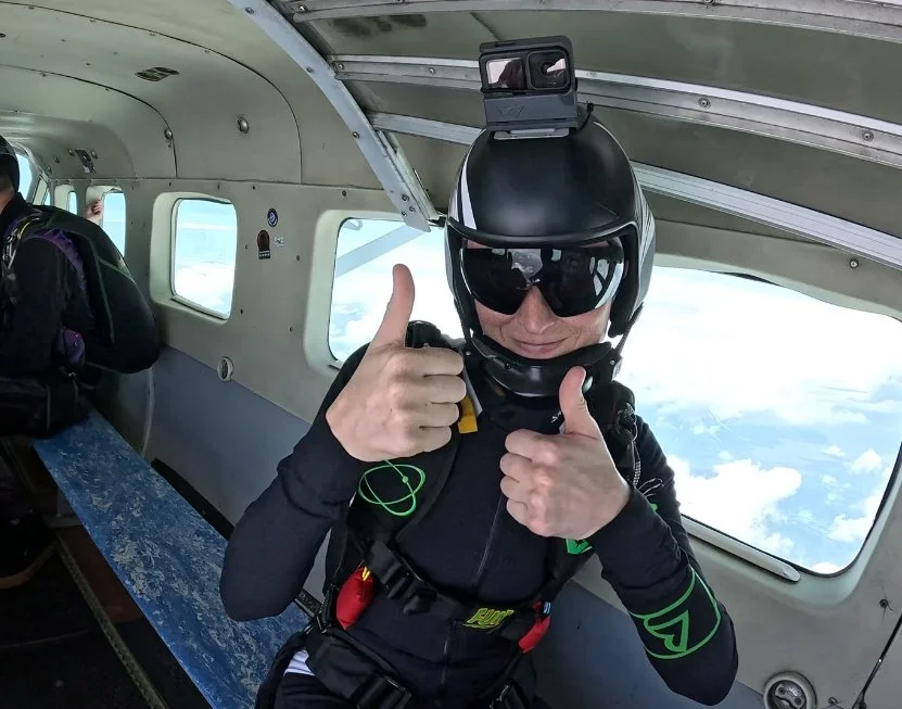 Skydiver inside airplane, wearing helmet and goggles, giving thumbs up.