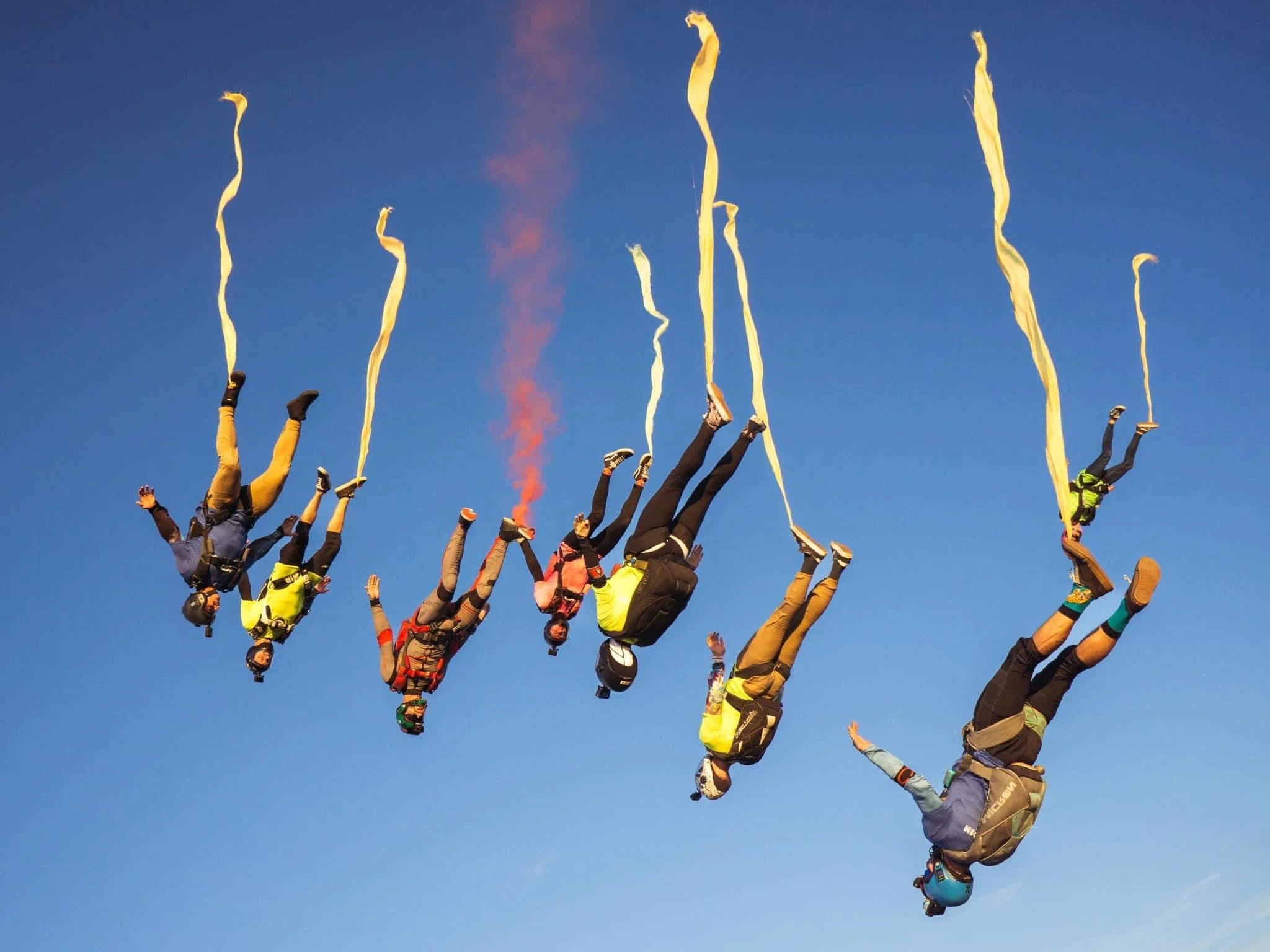 A group of skydivers in free fall, descending against a clear blue sky, holding yellow streamers and wearing colorful jumpsuits and helmets.