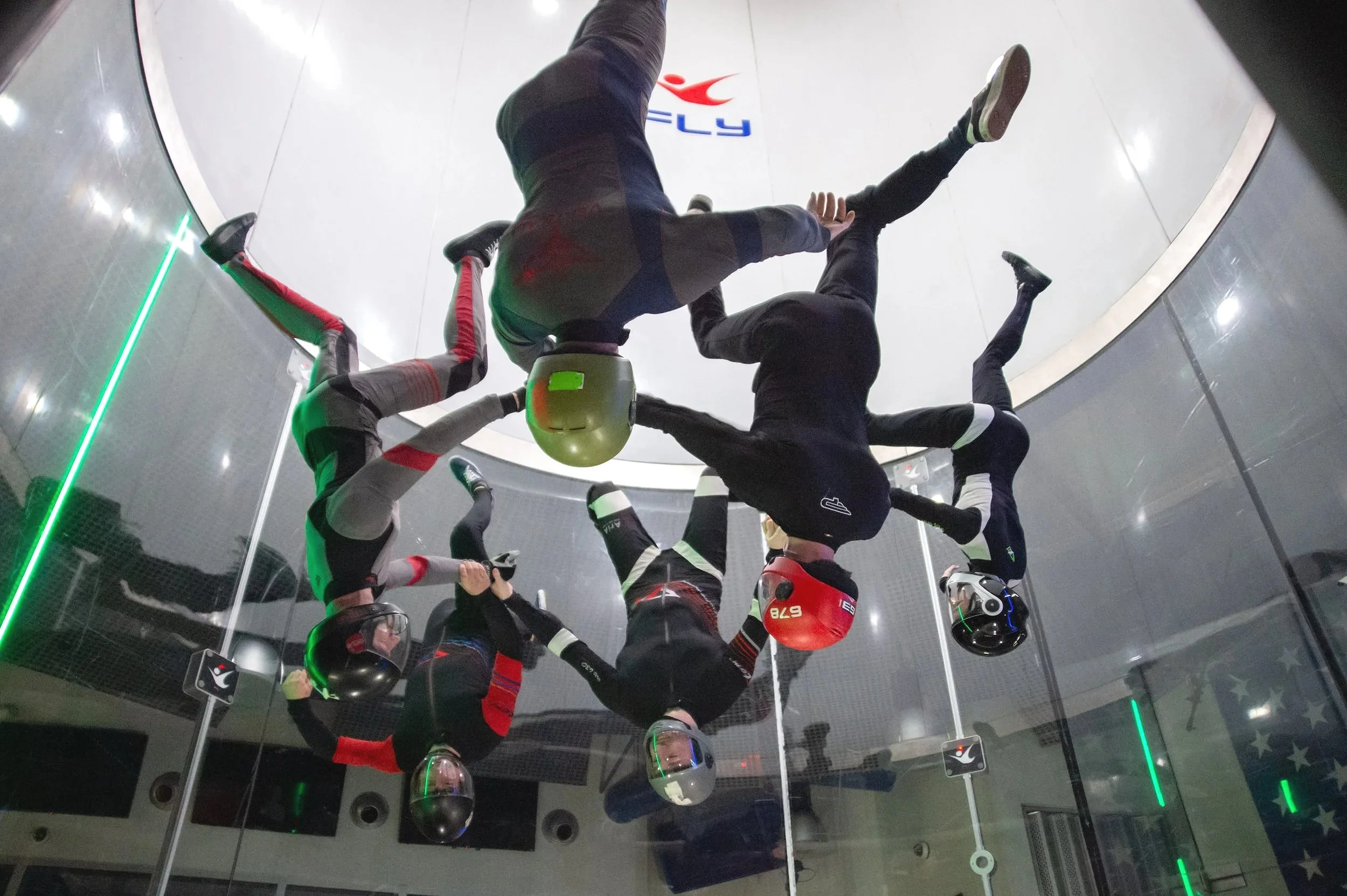 Group of people in a wind tunnel wearing helmets and suits, practicing indoor skydiving.