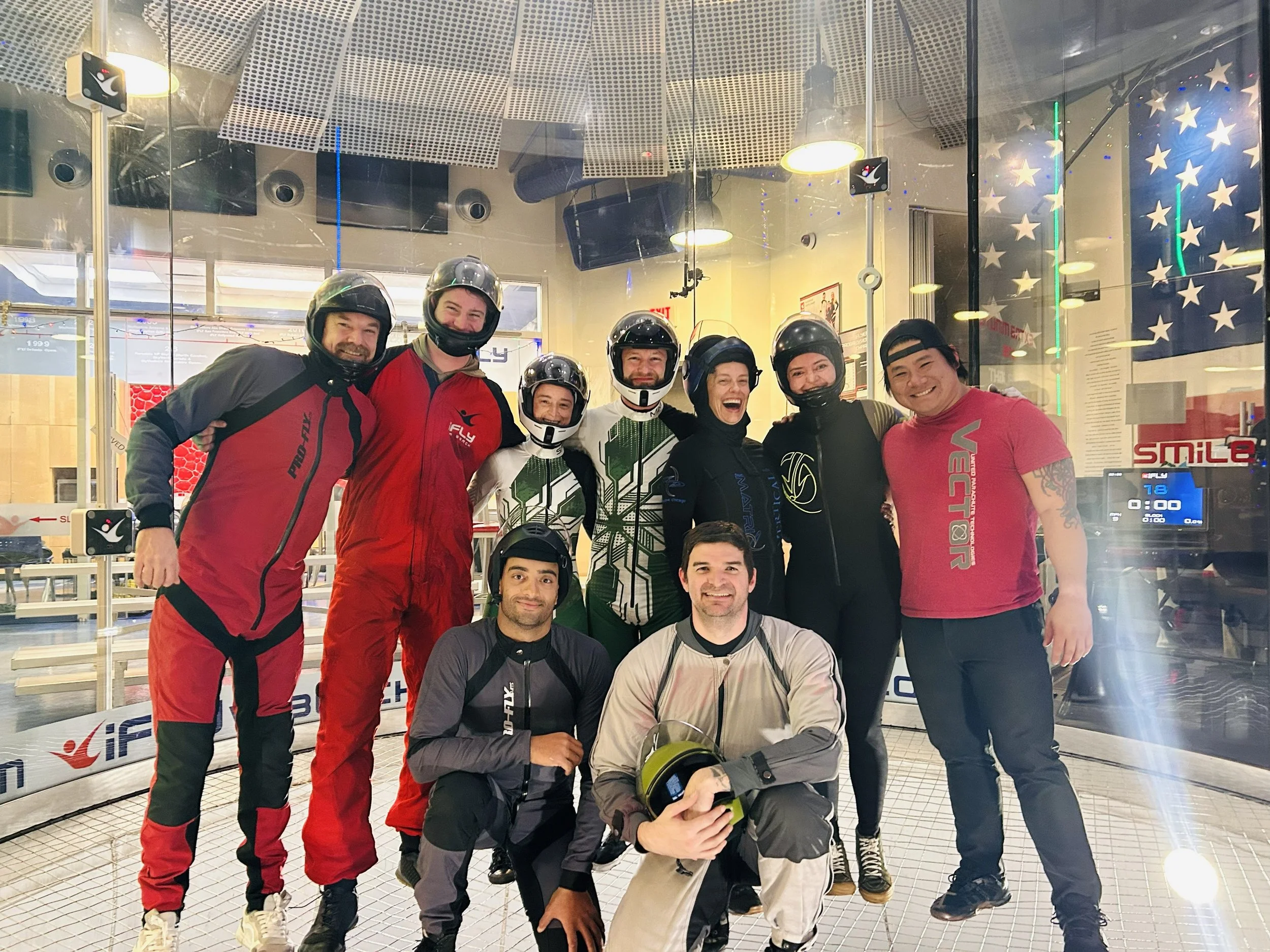 A group of eight people wearing motorcycle helmets and gear pose inside an indoor skydiving facility, with a glass wind tunnel and American flag design in the background.
