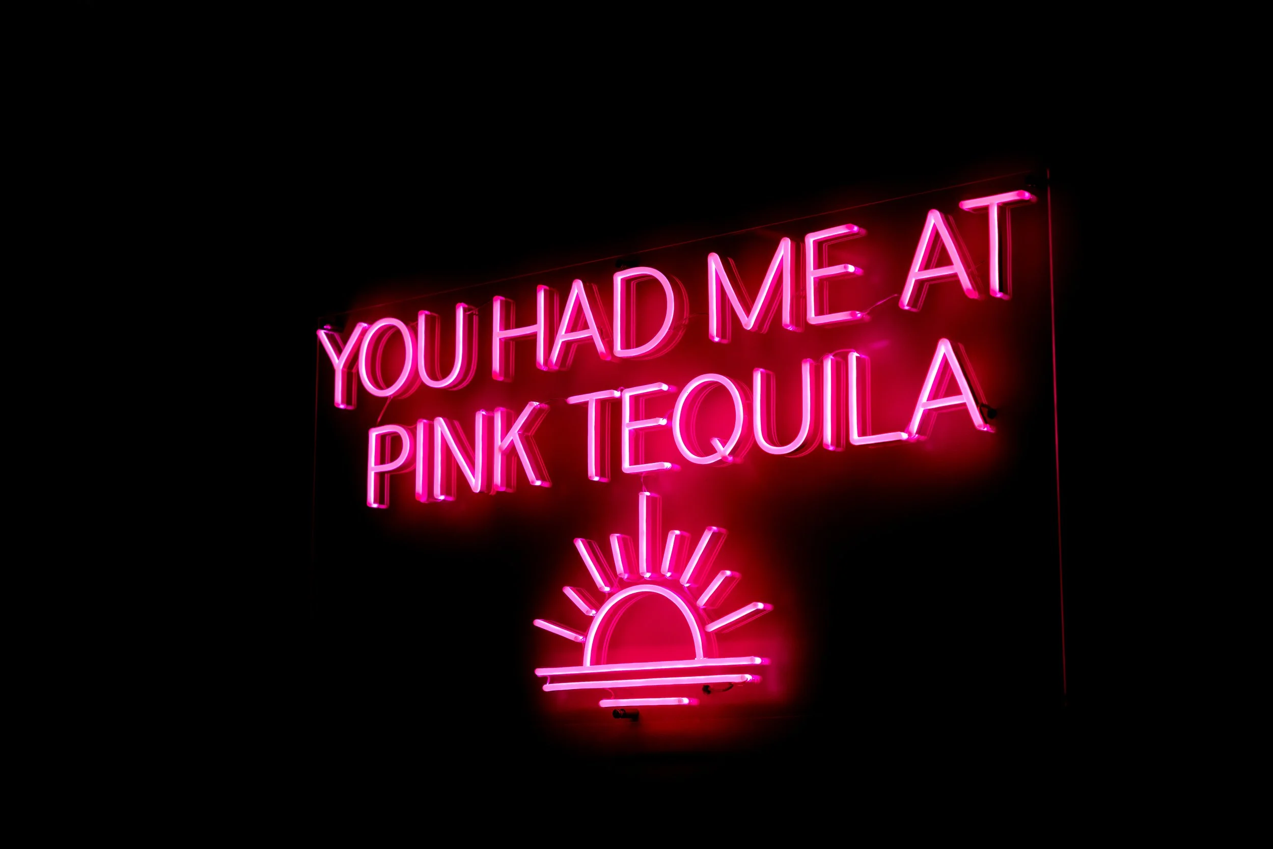 Neon sign reading "You had me at pink tequila" with a stylized pink sun and rays below the text, against a black background.
