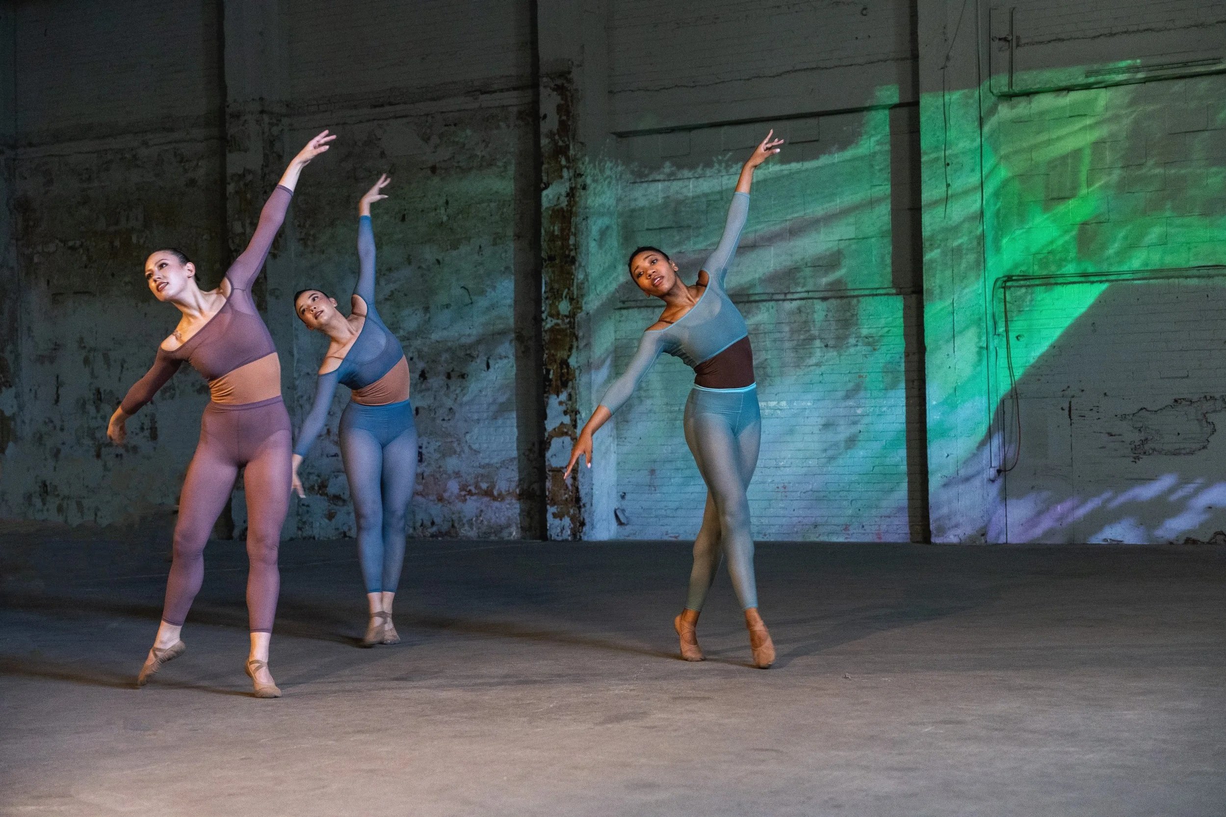 Four ballet dancers practicing in an industrial-style studio, wearing fitted dance attire with neutral tones, capturing a graceful pose with arms raised and tilted heads.