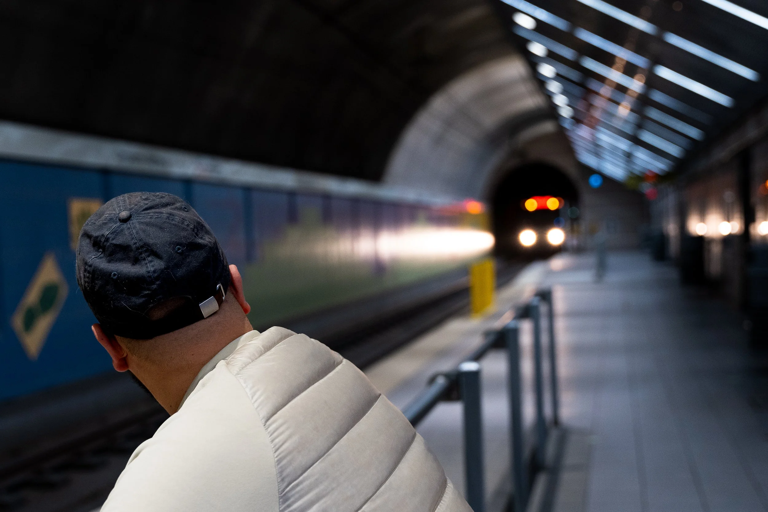 A person wearing a black cap and a cream-colored puffer jacket waiting on a subway platform as a train approaches in the background.