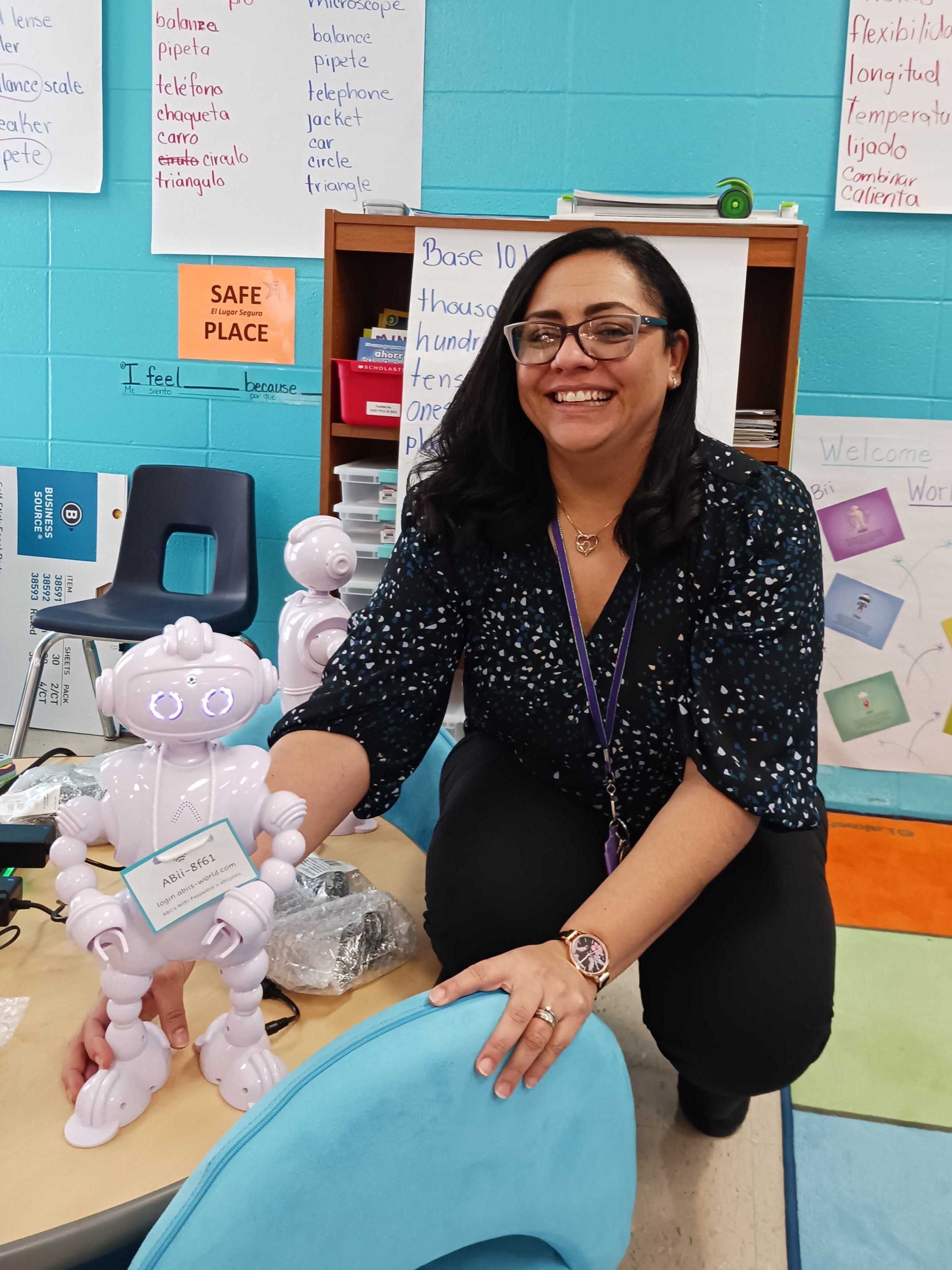 A woman smiling in a classroom, kneeling beside a table with a white robot toy. The classroom has educational posters and colorful decor.