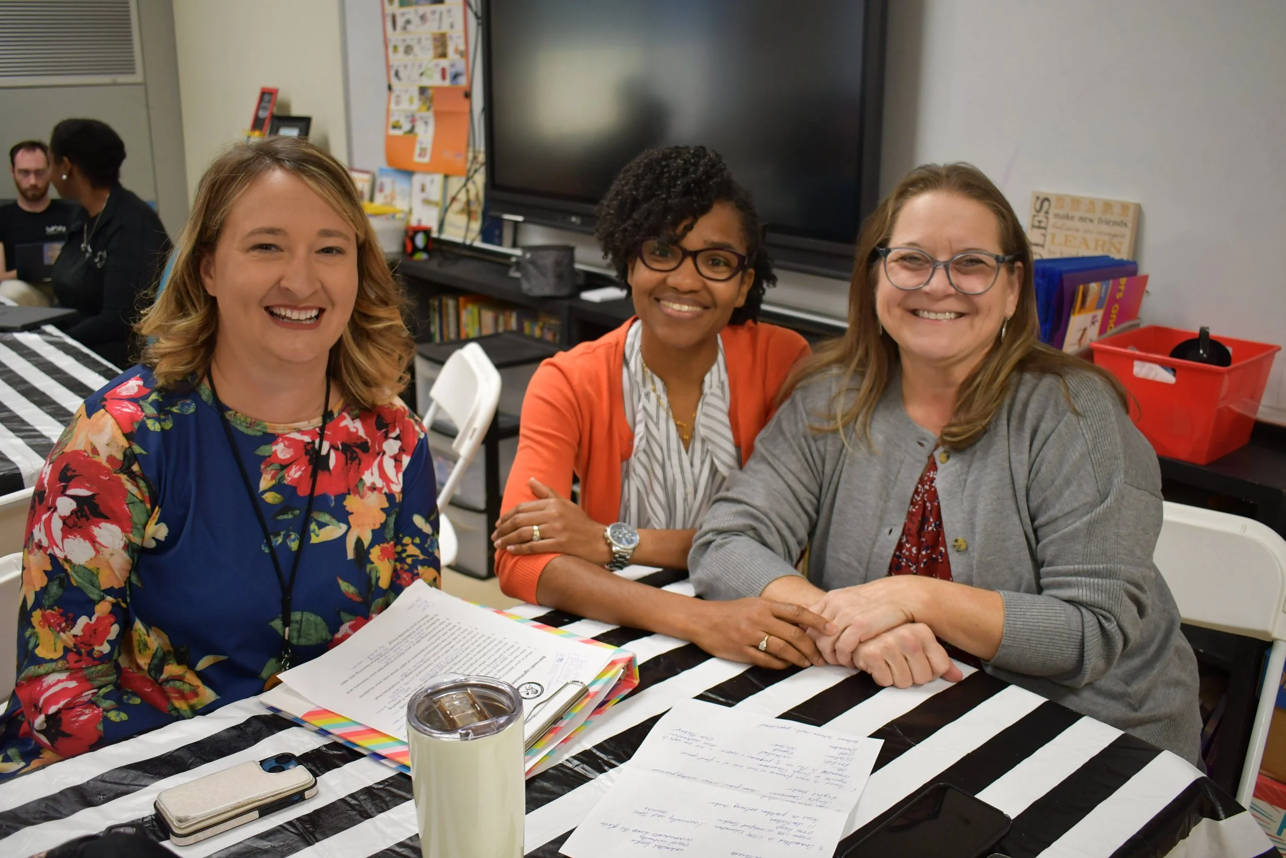 Three women smiling at a table with documents and a tumbler in a classroom setting.