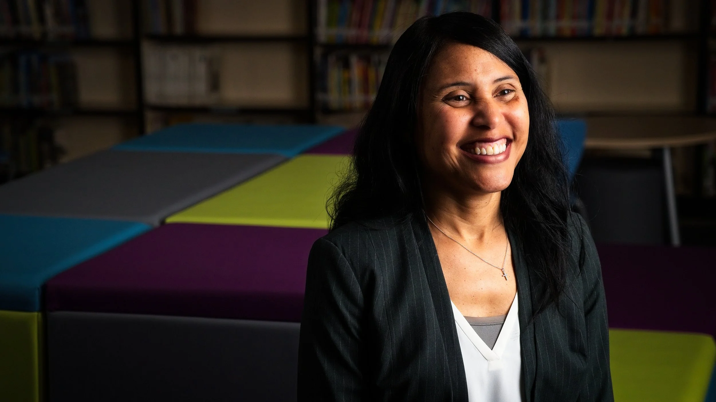 A woman smiling, sitting indoors with colorful background blocks and bookshelves.