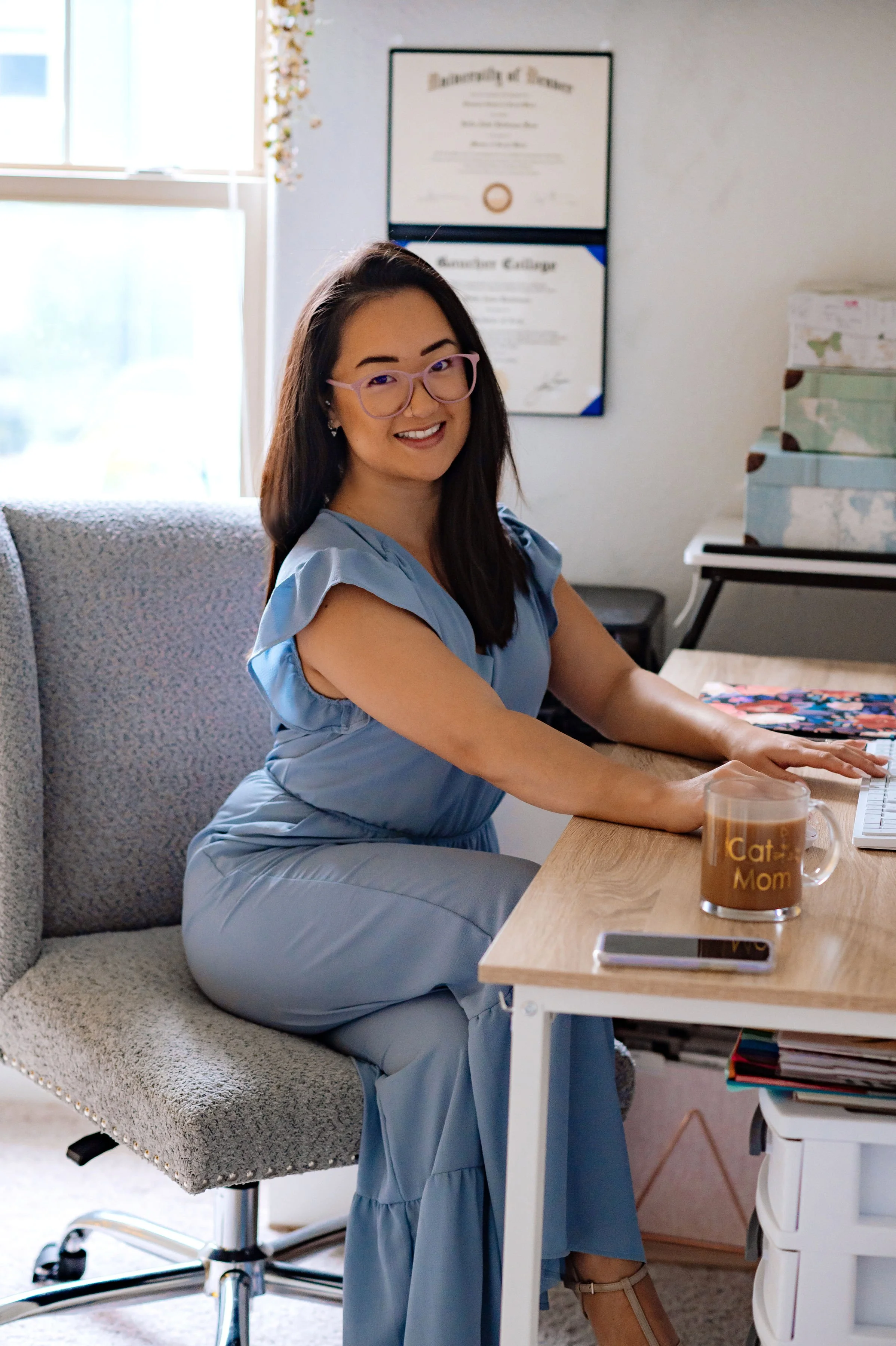 A woman sitting on a beige office chair at a desk, smiling, wearing glasses and a light blue dress, with a coffee mug that says 'Cat Mom' in front of her, and a smartphone on the desk. There are framed documents on the wall and stacked boxes in the background.