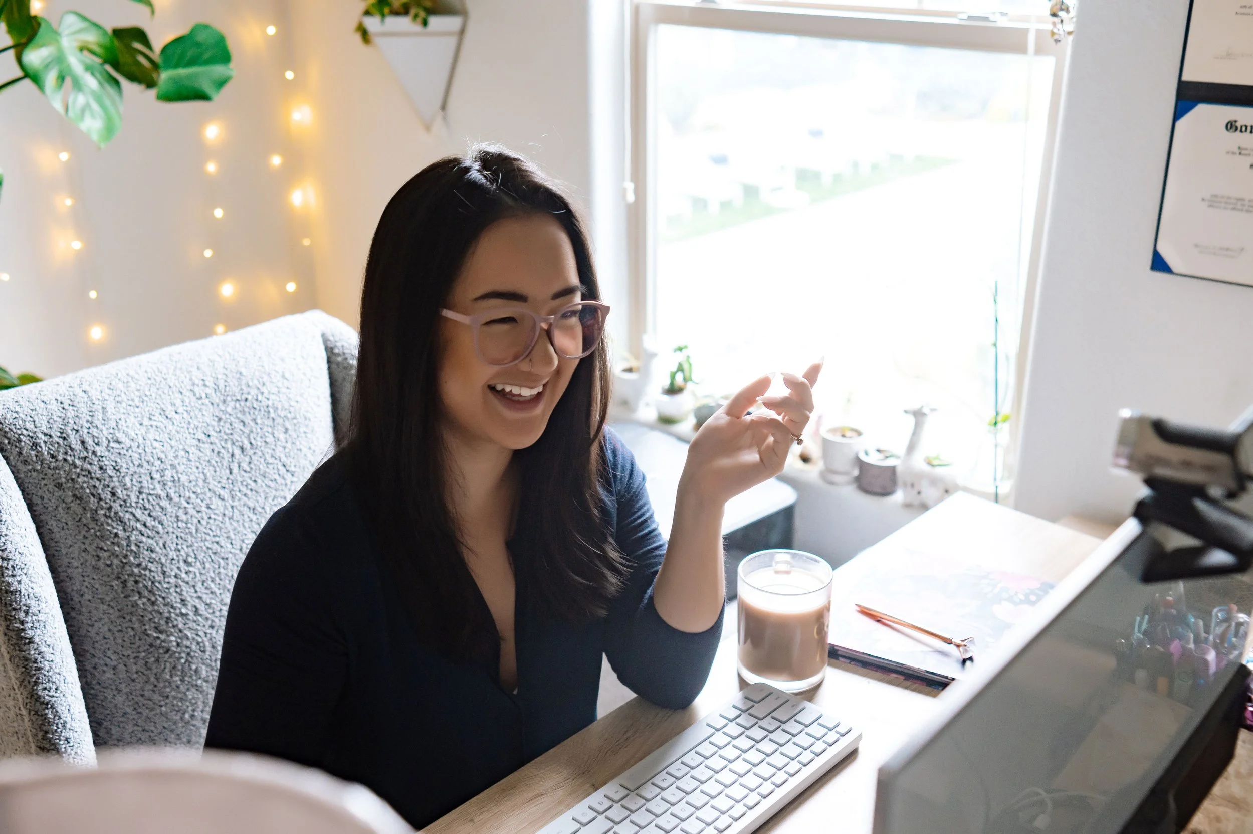 A woman with long dark hair and glasses smiling at her computer while sitting at a desk with a glass of iced coffee, in a bright room with a window, plants, and decorative string lights.