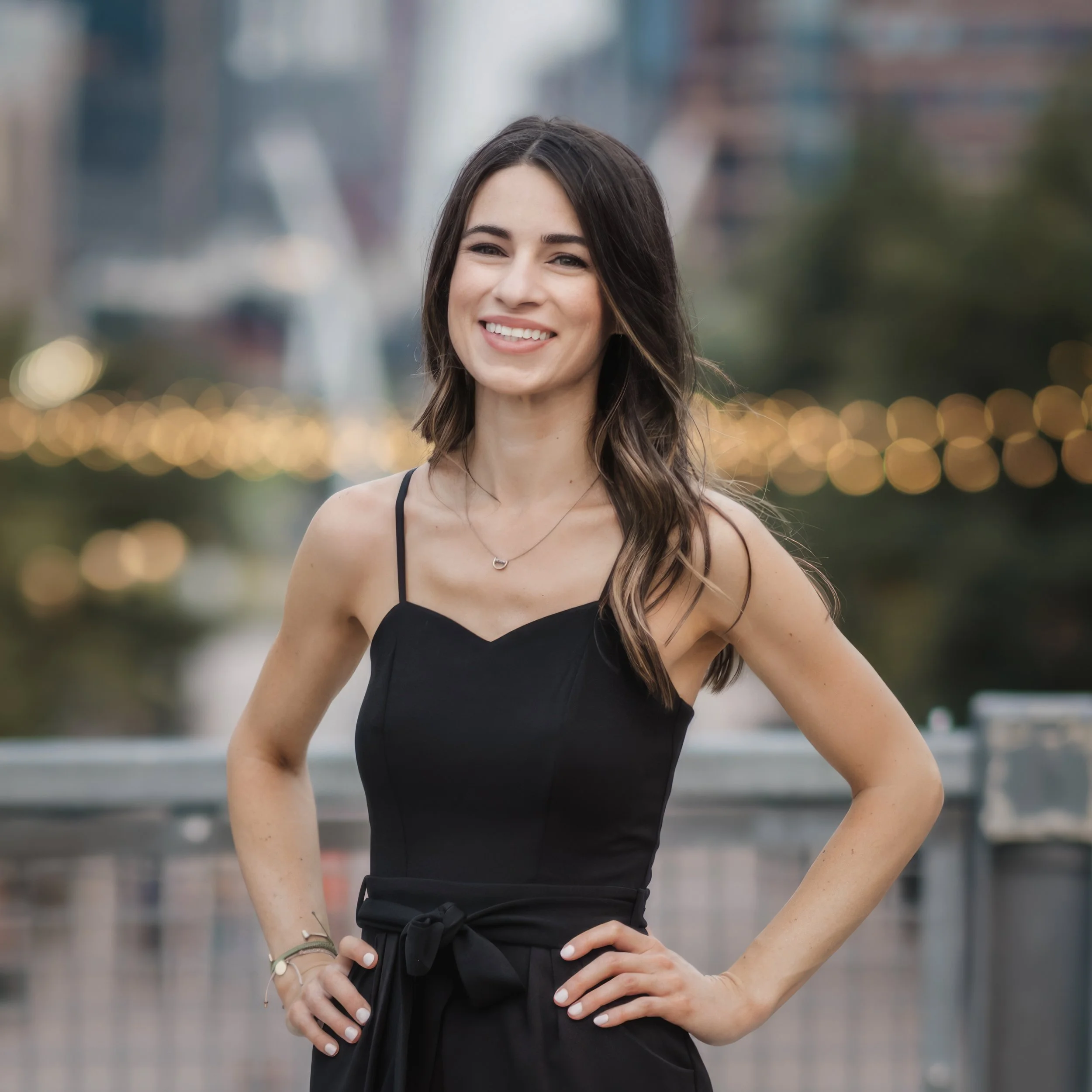 A woman with long brown hair, wearing a black sleeveless dress and a necklace, smiling outdoors in an urban setting during evening.
Colorado photographer