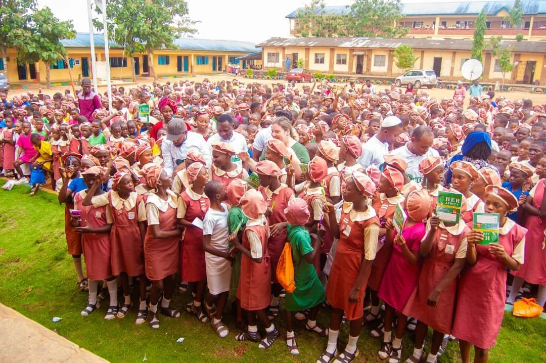 Large group of school children and teachers gather outside in a schoolyard with yellow buildings in the background, some children holding books and wearing school uniforms and hats.