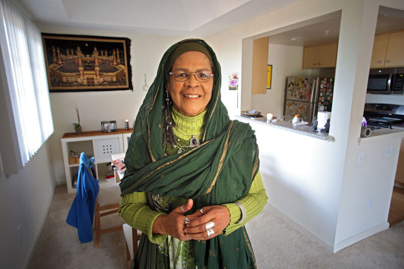  Dr. Amina Wadud in her home. Behind her at left is a wall hanging depicting the Kaaba in Mecca.    Photo Credit: Tom Levy. 