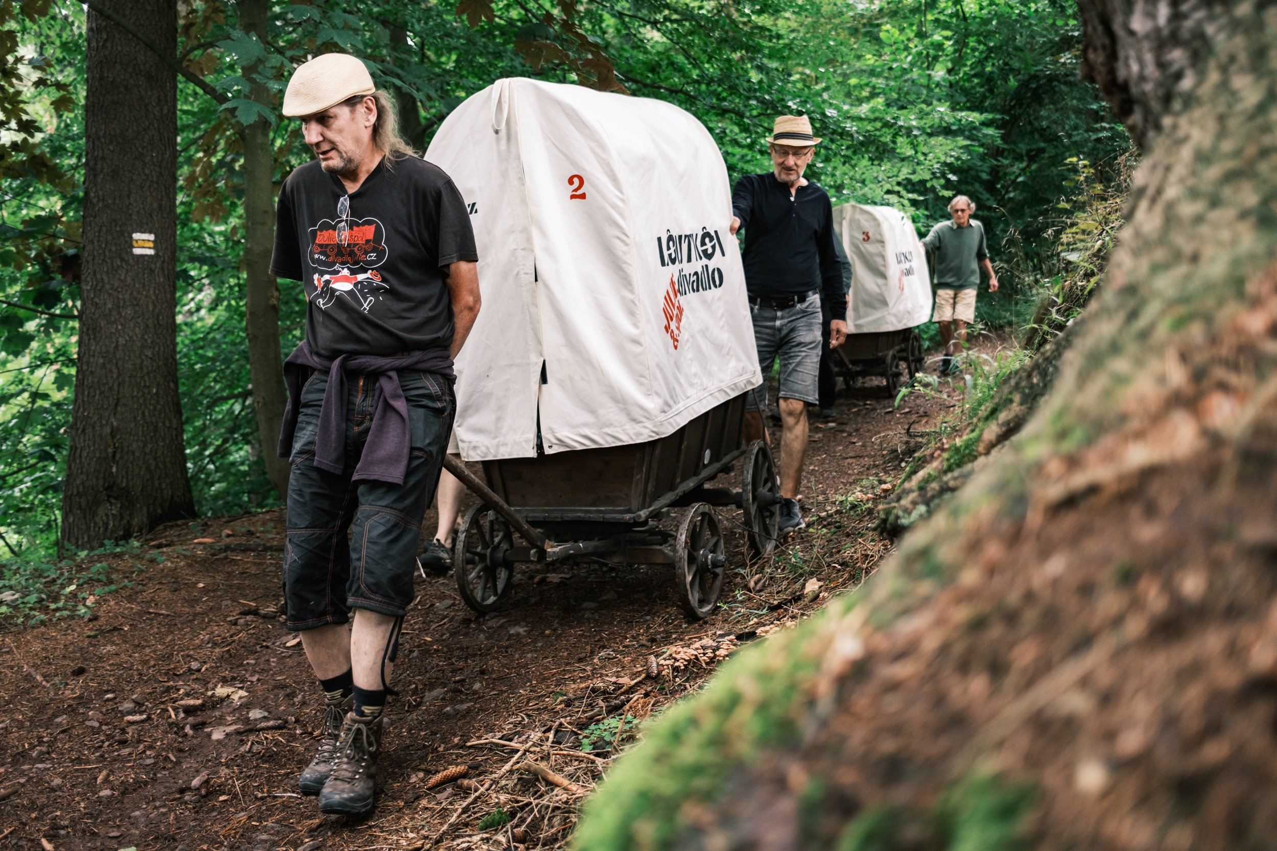 People walking in a forest pulling covered wagons labeled with text, wearing casual outdoor clothing.