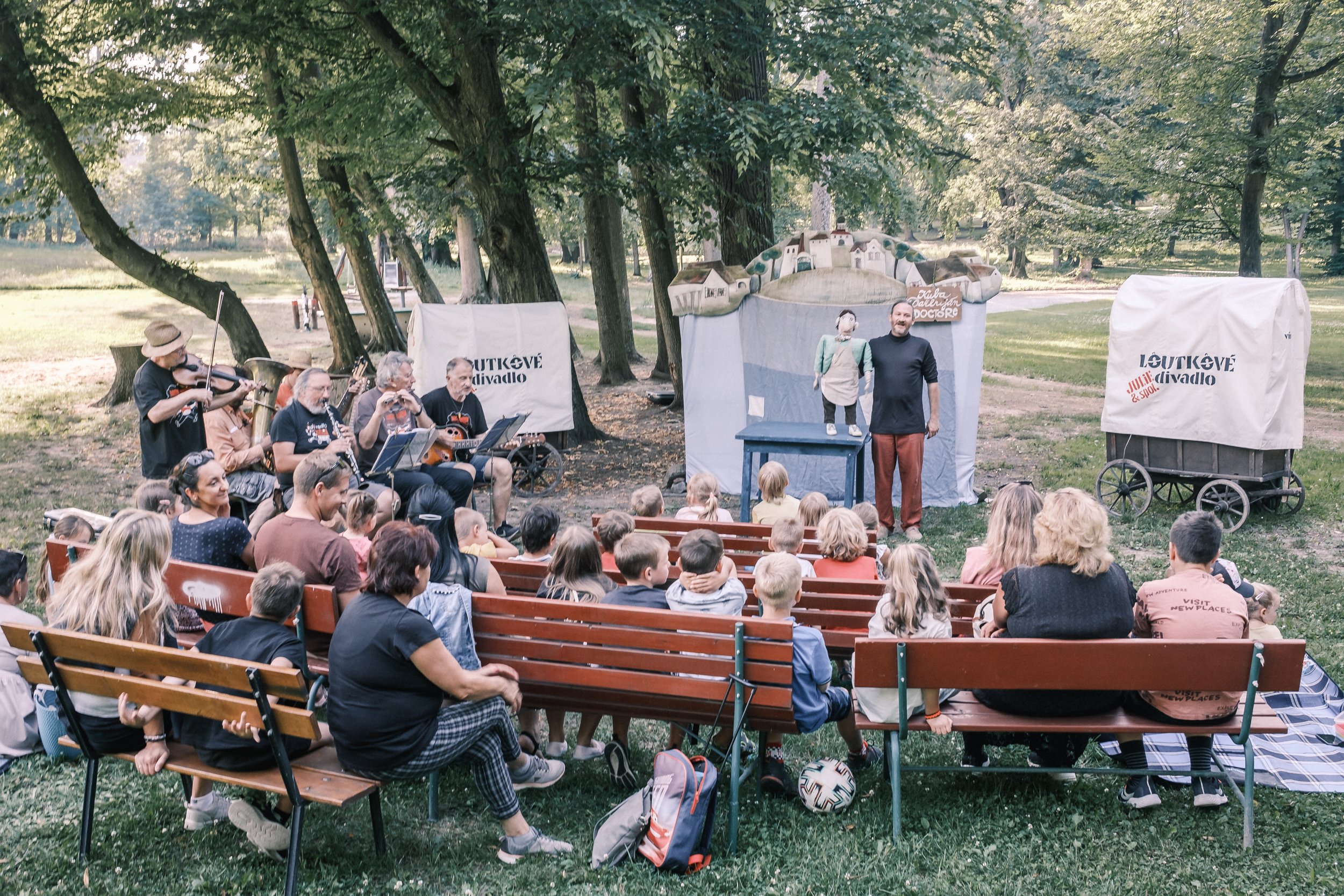 Outdoor puppet show in a park with an audience seated on benches watching a performer and a puppet, accompanied by musicians playing instruments. Trees and greenery in the background.