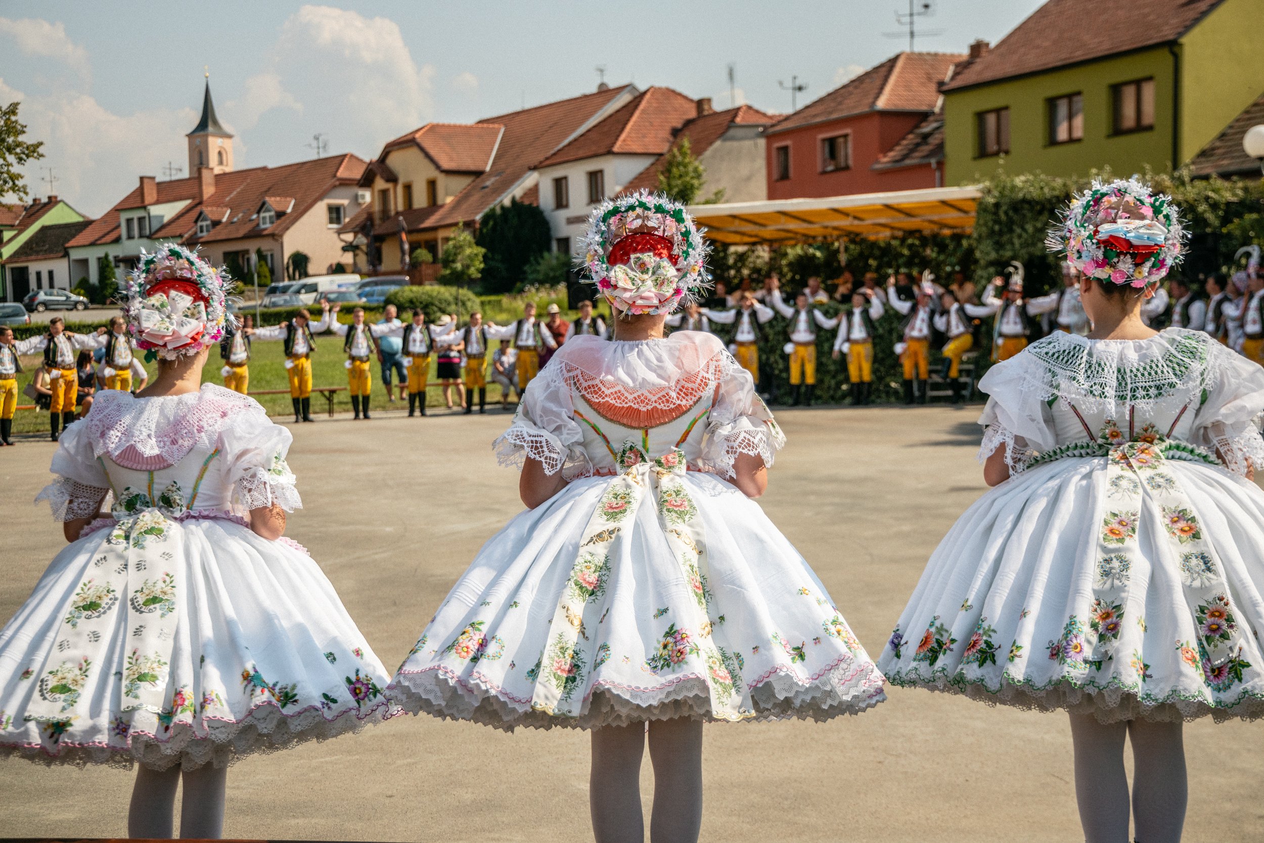 People in traditional Czech costumes during a festival, with three women in embroidered dresses and floral headpieces facing dancers and musicians in colorful attire, set in a village square.
