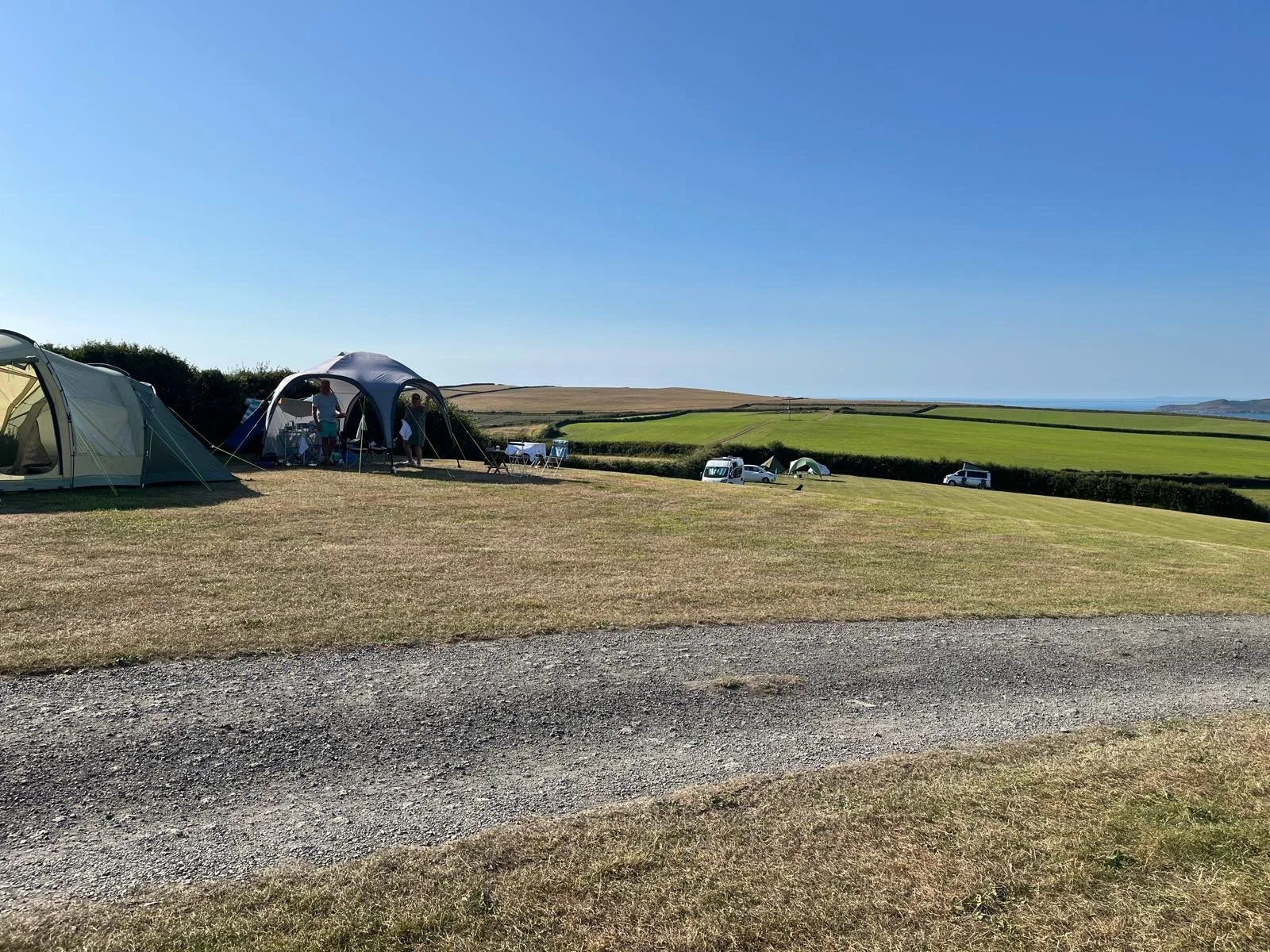 A campsite with multiple tents set up on a grassy field under a clear blue sky, with cars parked in the background and rolling hills in the distance.