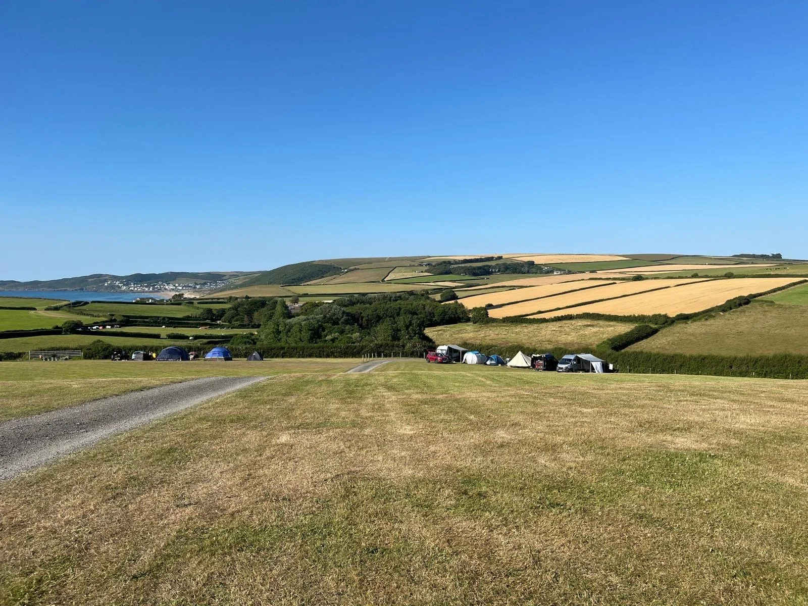 A panoramic view of a countryside with camping tents and vehicles, rolling green and yellow fields, and distant hills under a blue sky.