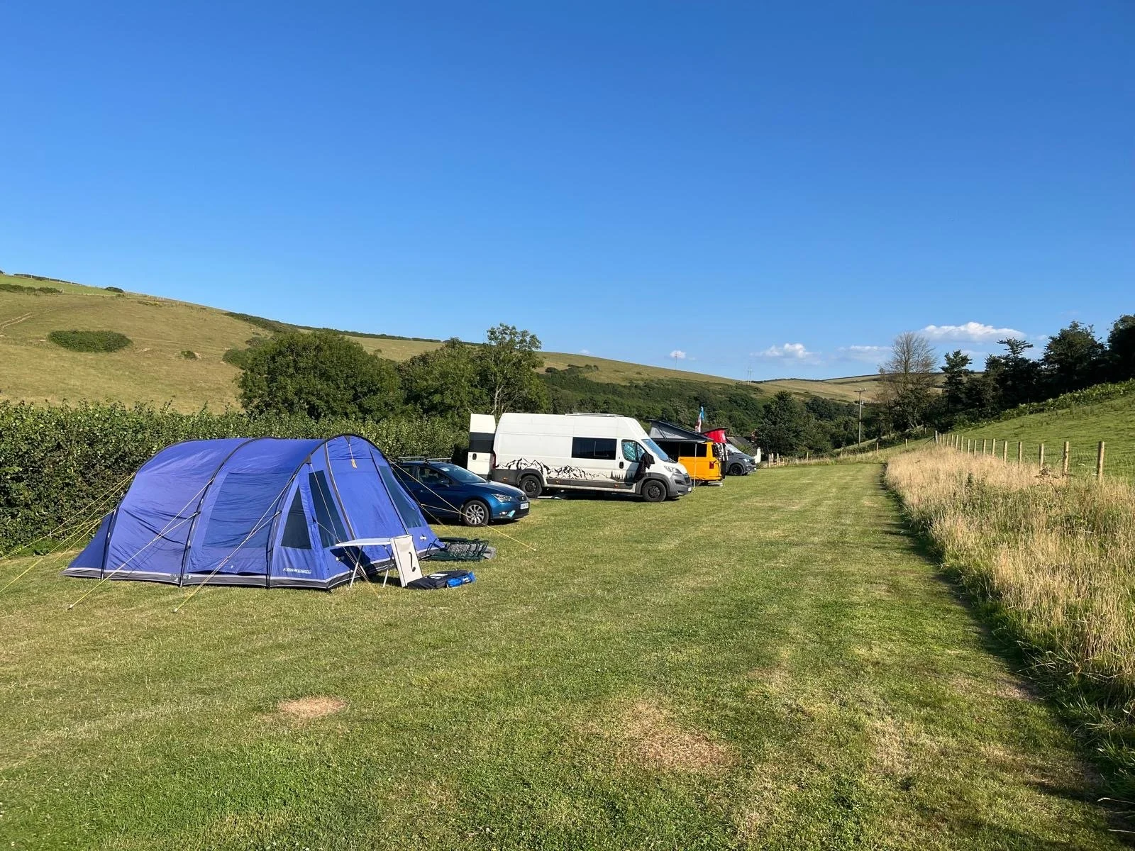 A daytime outdoor camping scene with a blue tent on a grassy field, several parked cars, including a white van, and a yellow camper van, surrounded by rolling green hills and a clear blue sky.