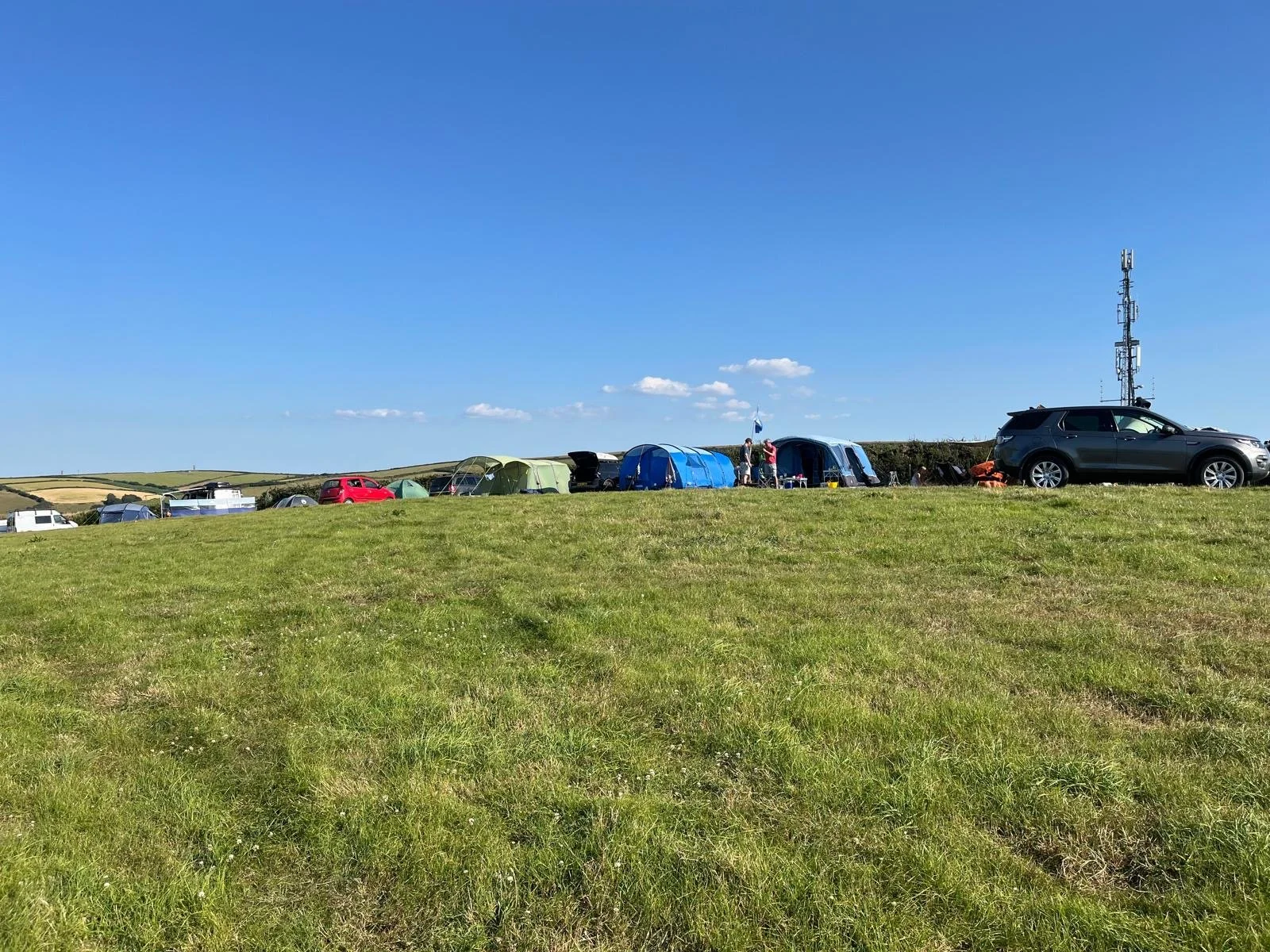 A grassy hill with various tents, cars, and people camping under a clear blue sky.