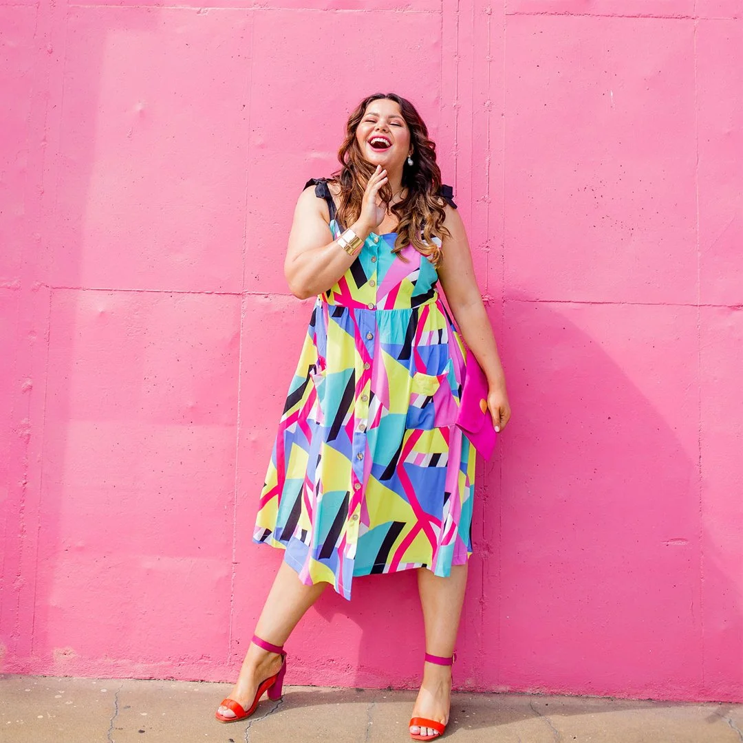 A woman wearing a colorful, patterned dress with pink, blue, yellow, and black, standing against a pink wall, smiling and playful.