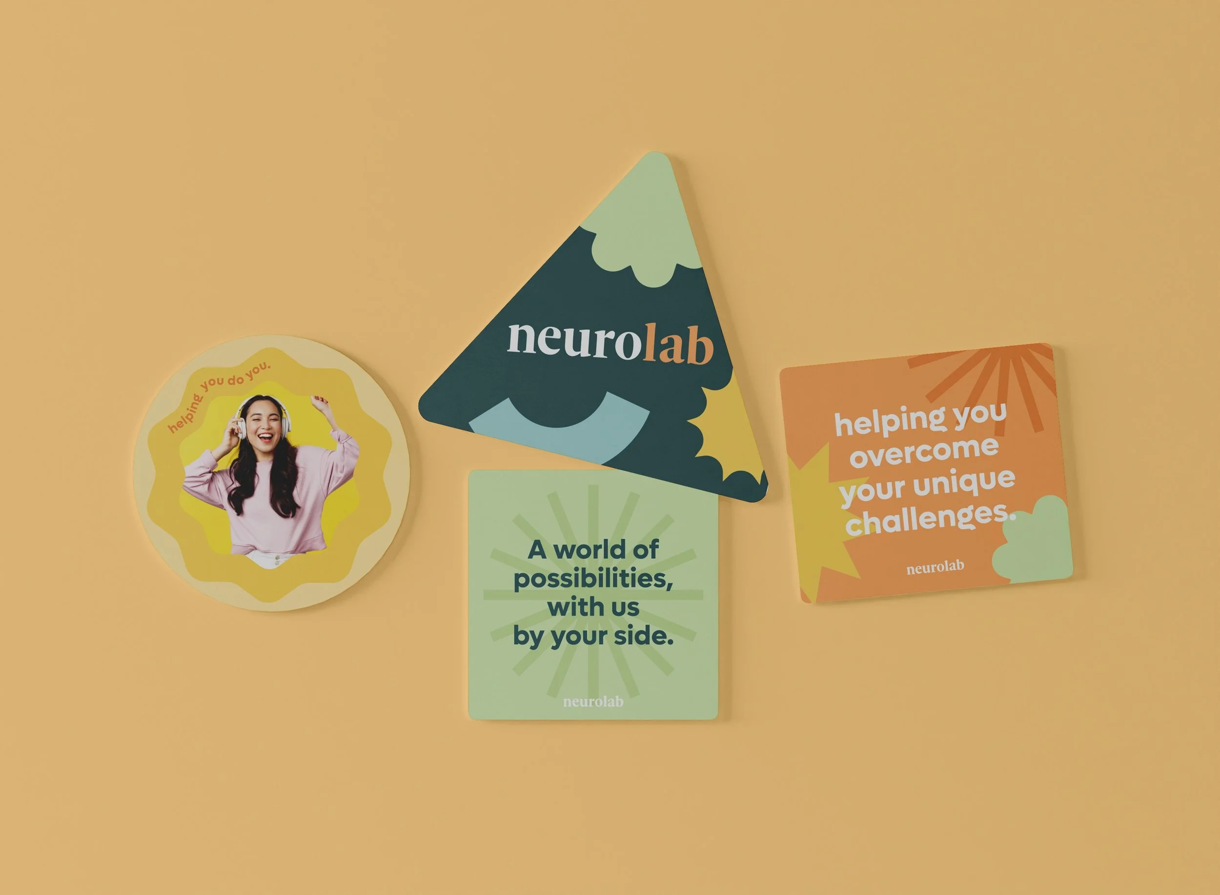 Four colorful promotional cards for NeuroLab on a peach background. The cards include a round card with a woman with headphones and the text "helping you do you." A dark green triangle card with "neuro lab" written on it. A rectangular green card with the text "A world of possibilities with us by your side." A square orange card with the text "helping you overcome your unique challenges."