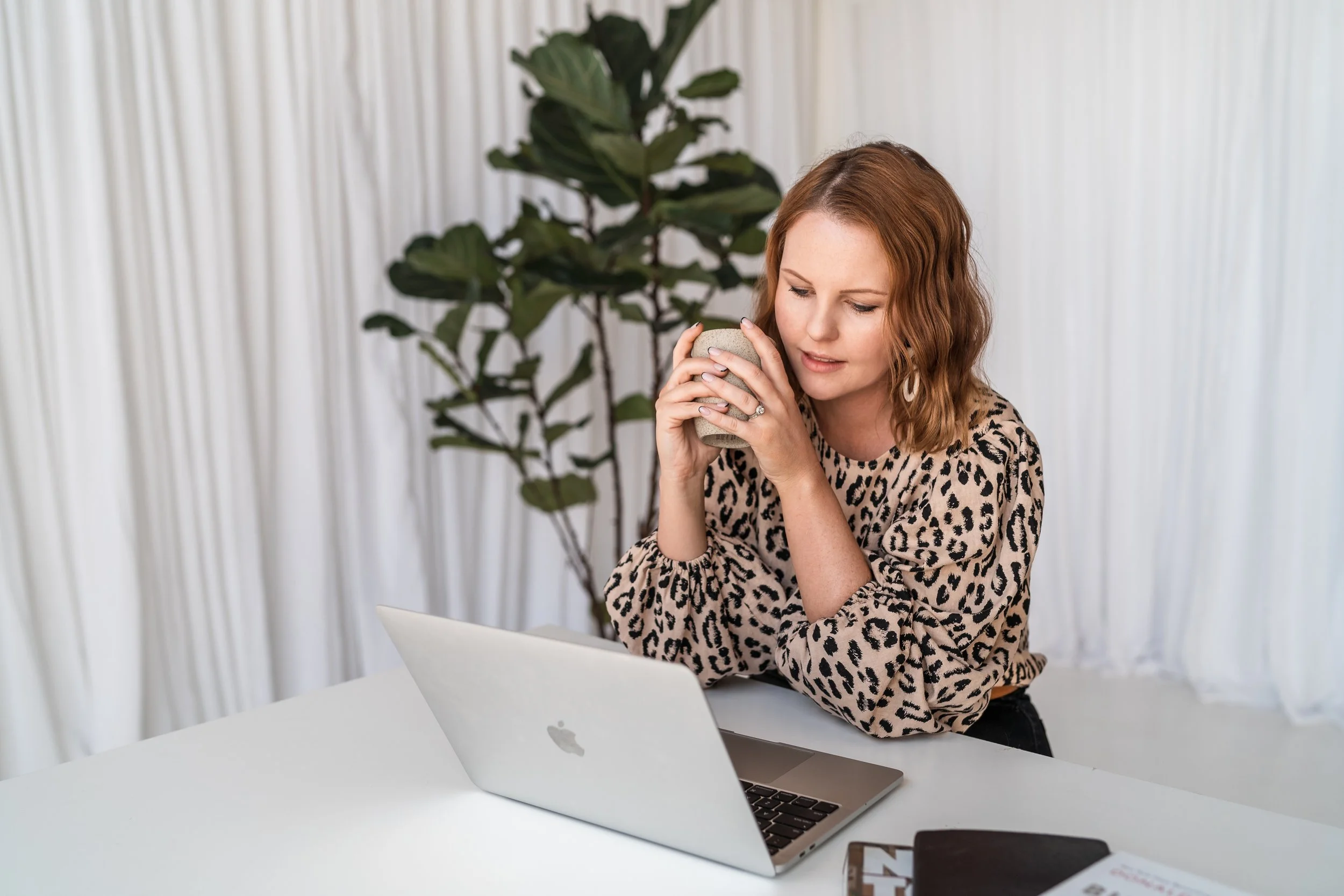 A woman with wavy, shoulder-length red hair wearing a leopard print blouse, holding a mug close to her face while sitting at a white desk with an open silver MacBook. Behind her is a large green plant and white curtains.
