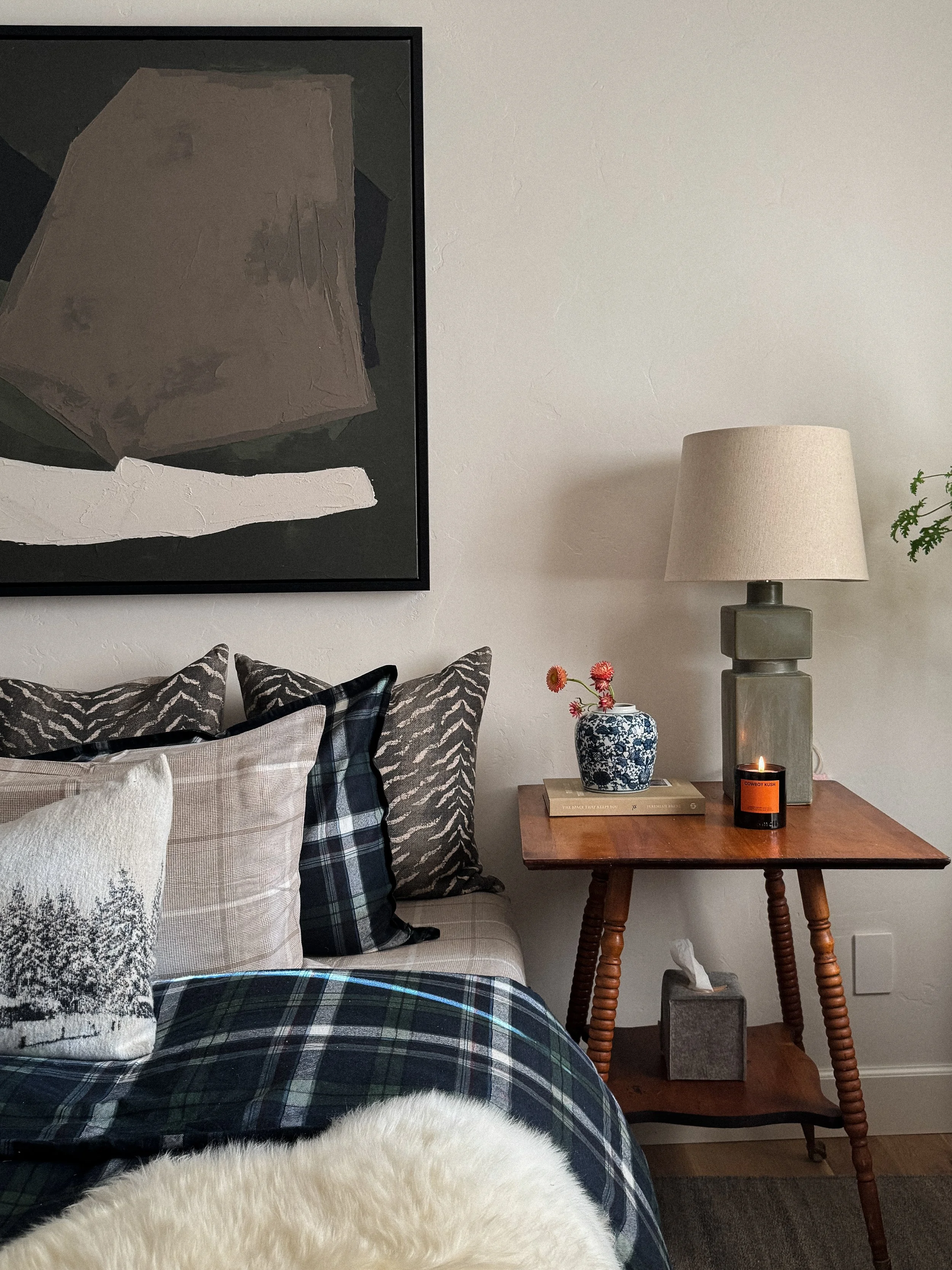 A bedroom corner with a bed and pillows, a nightstand with a decorative vase holding flowers, a book, a lit candle, and a lamp, with an abstract painting above.