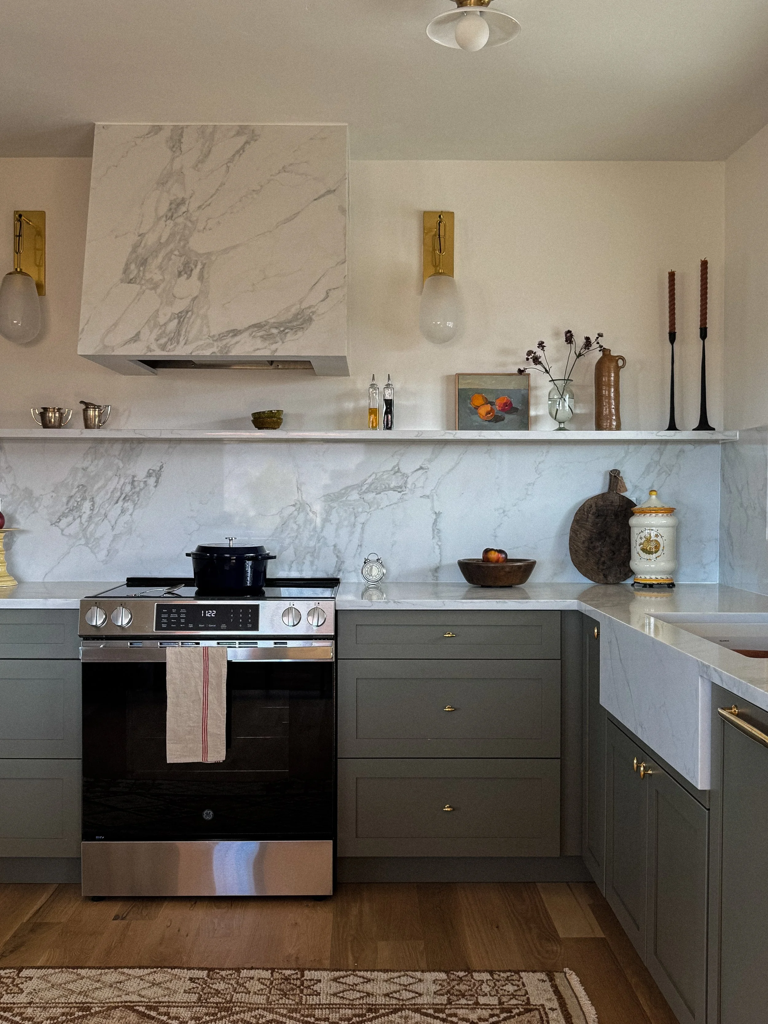 Mountain modern kitchen design in Steamboat Springs, Colorado, featuring green sage cabinets, unlacquered brass hardware, wall sconces & a marble countertop, shelf & range hood detail.