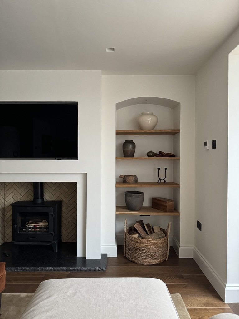 Living room with a television, wood-burning stove, built-in shelves with decorative jars and objects, wicker basket with firewood, and part of a beige upholstered armchair.