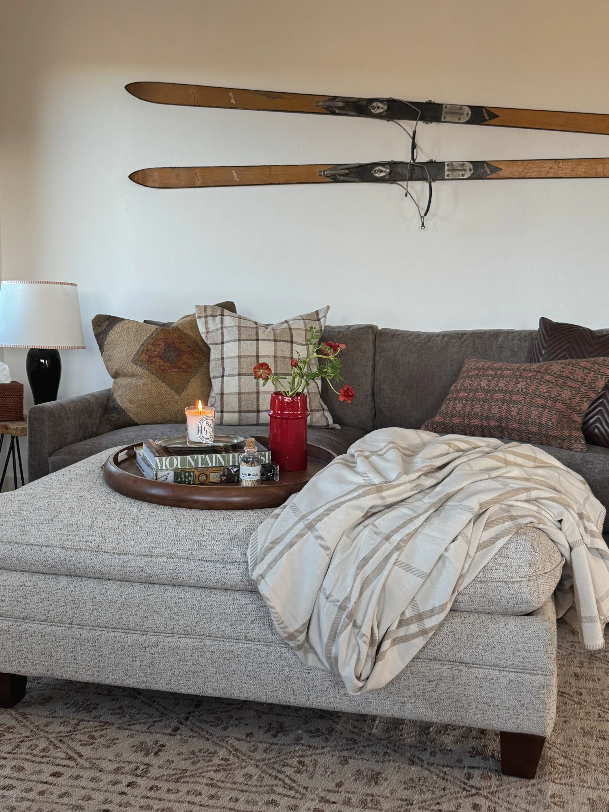 A cozy and refined mountain living room in Steamboat Springs, Colorado, featuring a gray sectional, layered throw pillows in plaid and vintage textiles, and a large upholstered ottoman styled with a wood tray, candle, and red vase.