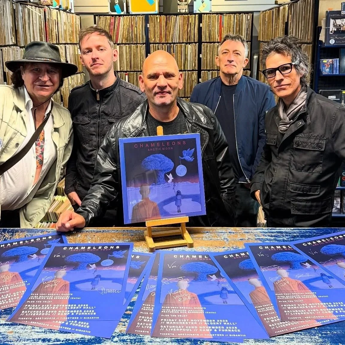 A group of five people standing behind a table with promotional flyers for Chameleons' Arctic Moon Tour. The person in the center is holding a framed poster of the tour. They are in a store with shelves of vinyl records in the background.