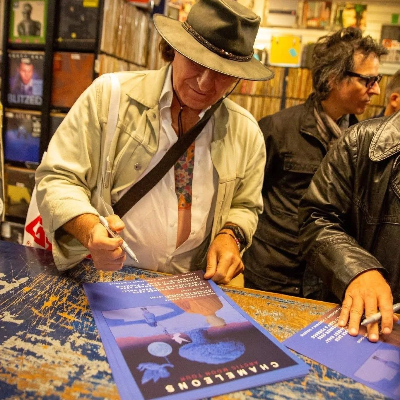 A man wearing a hat and light-colored jacket signs a poster at a book signing event, with other people around him and shelves of books in the background.
