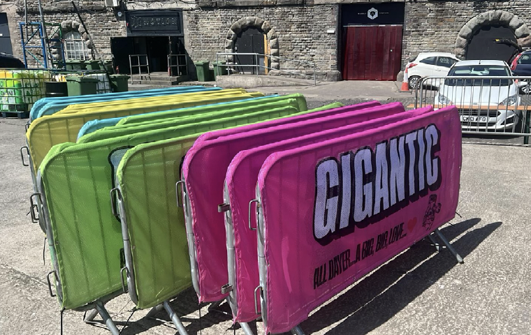 Colorful barriers in pink, green, and blue, with the pink barrier prominently displaying the word "GIGANTIC" and the slogan "ALL DAY, ALL LOVE." parked outdoors near a brick building, with cars and metal barricades in the background.