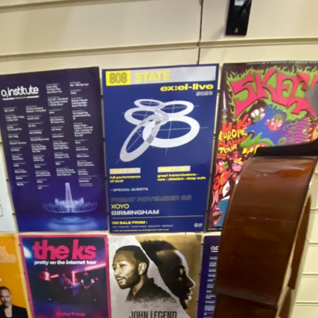 Close-up of a bulletin board with various colorful posters, including a music event with a blue background, a graffiti-style poster, and a pink and black poster for a tour, partially covered by a wooden object.