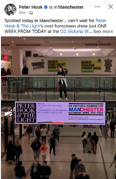 A woman standing in an airport terminal in front of a digital display board advertising Peter Hook & The Light concert in Manchester. The background shows other travelers and large posters on the wall.