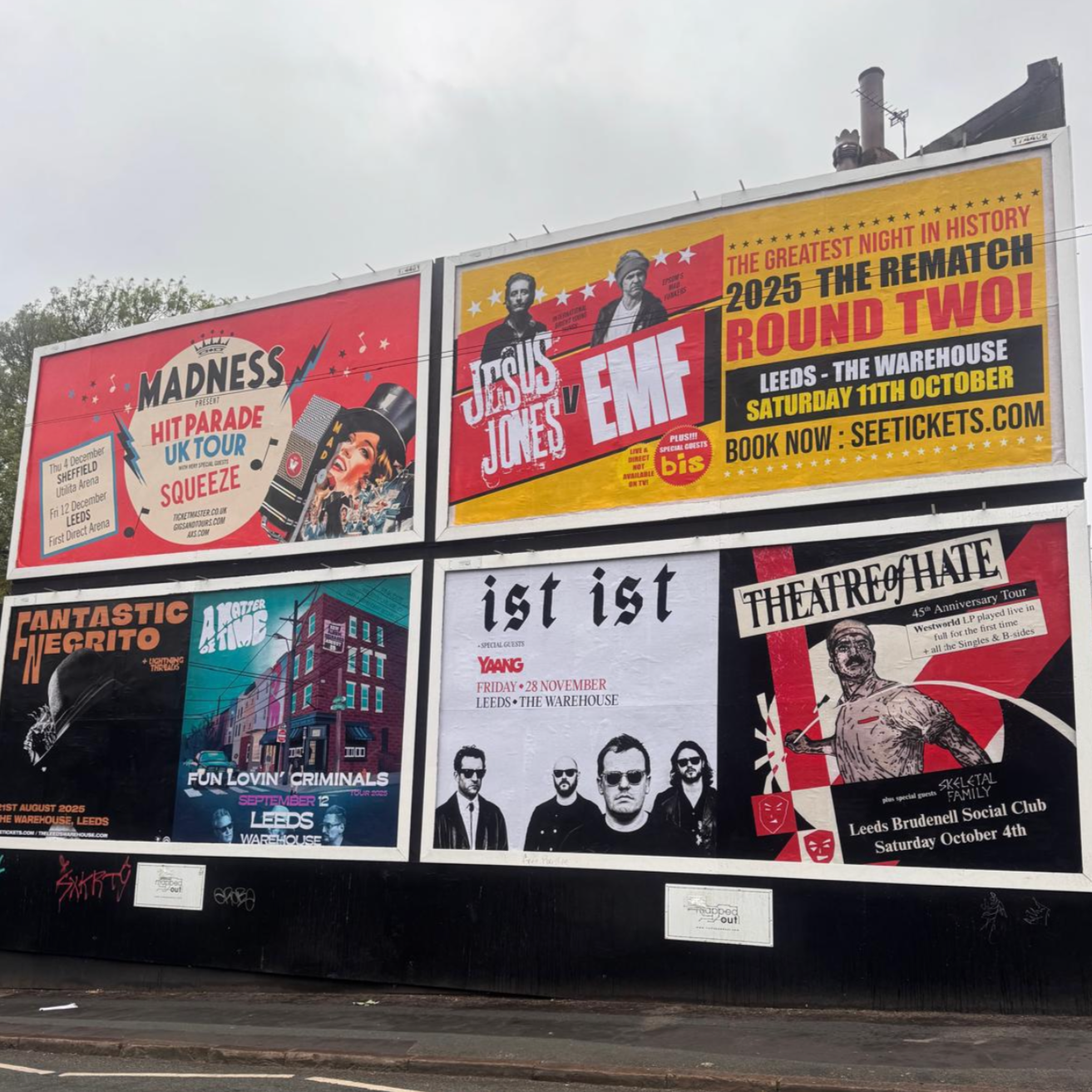 Billboard advertisements for various music and theater events, including hit parade UK tour, Jesus vs EMF, spectacular alien show, fun-loving criminals, and a theatre of hate tour, displayed outdoors under an overcast sky.