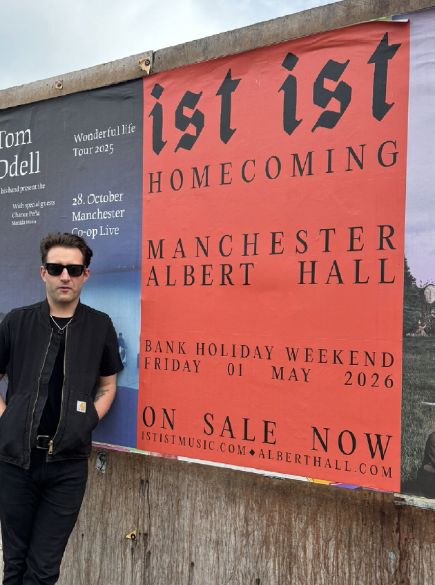 A young man in sunglasses and a black vest standing next to a large red and black promotional poster for the band "ist ist" advertising their homecoming event at Manchester Albert Hall on May 1, 2026, during a bank holiday weekend.