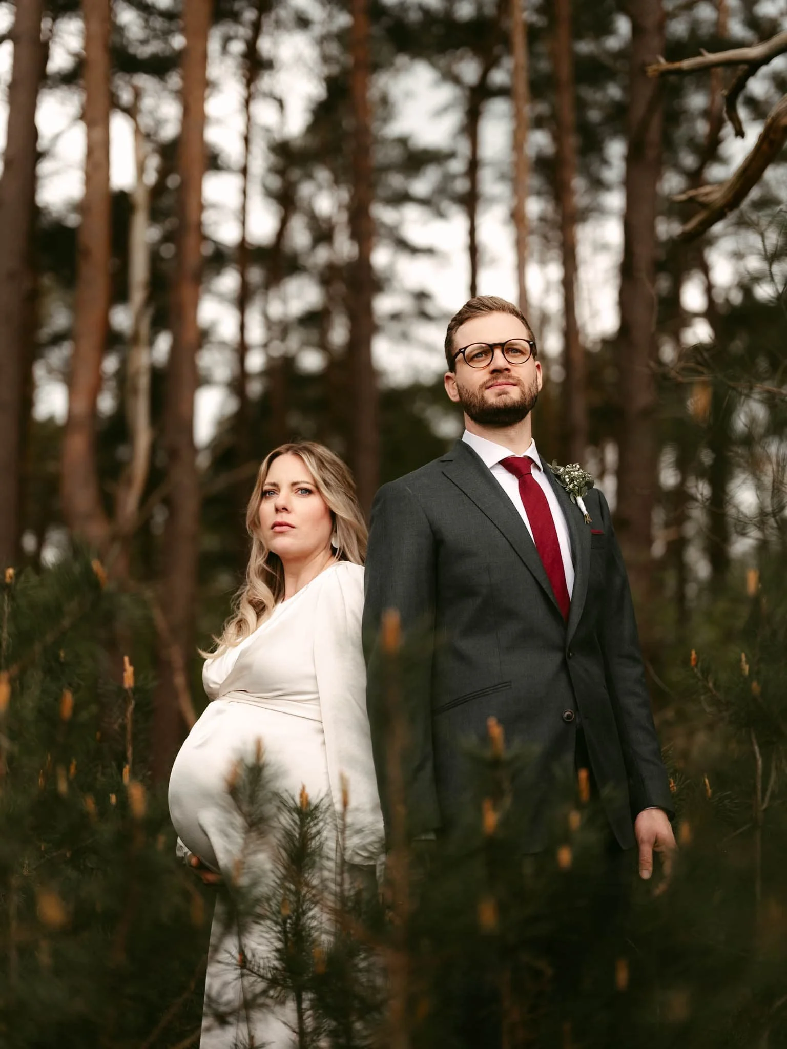 Dieses eindrucksvolle Foto, aufgenommen vom Hochzeitsfotografen Sturmsucht vom Ammersee, zeigt ein selbstbewusstes Paar in einem stillen Moment im Wald, wobei die werdende Mutter in einem weißen Kleid ihre Schwangerschaft stolz präsentiert