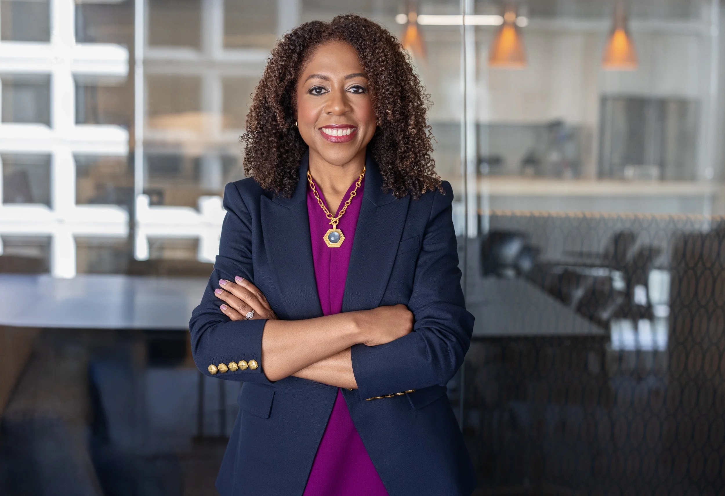 Tjada D’Oyen McKenna, CEO of Mercy Corps, standing confidently with arms crossed in a glass-walled office, wearing a navy blazer and magenta dress.