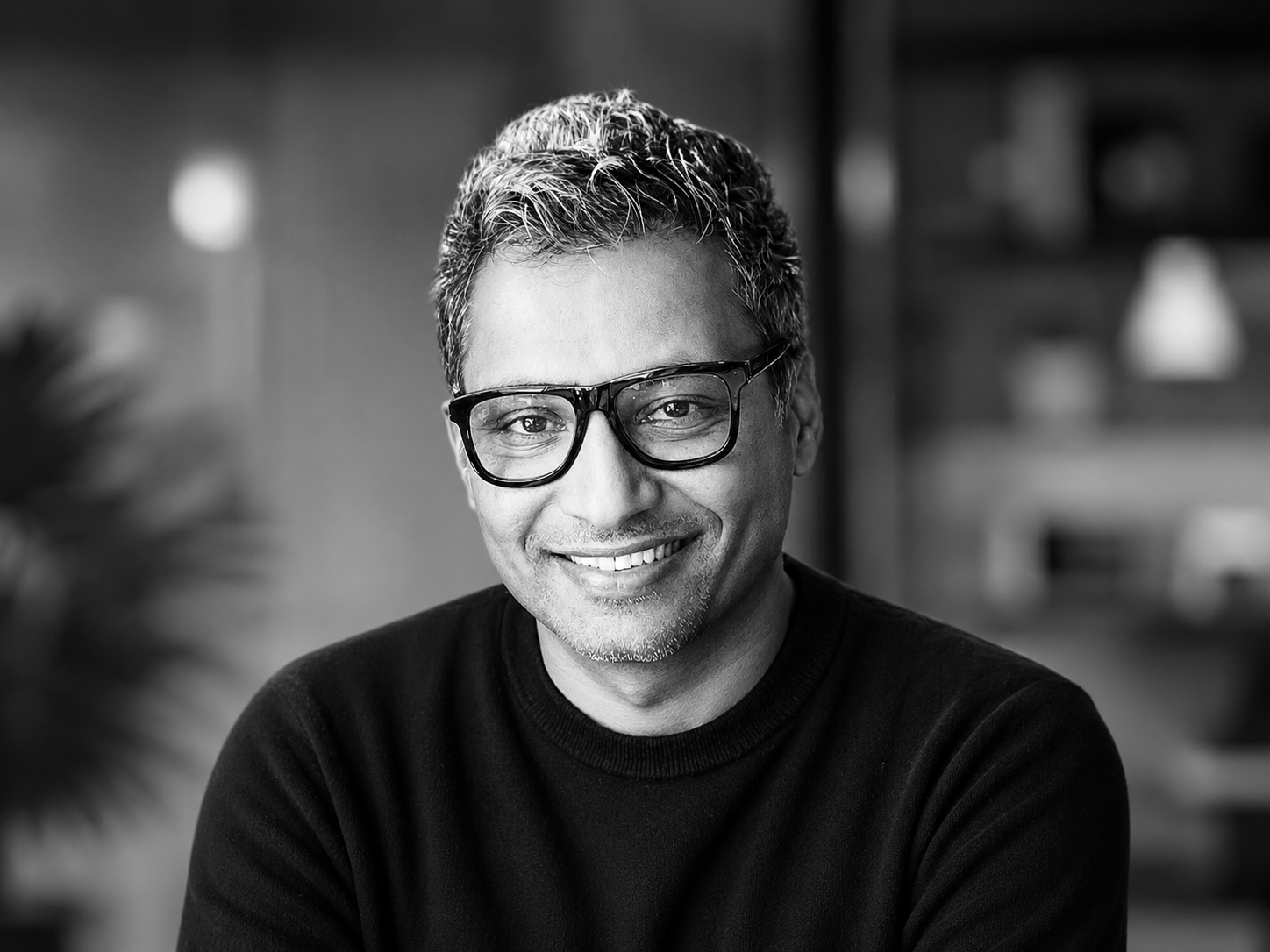 A portrait of Babith Bhoopalan in black and white. He is wearing black rimmed glasses a black top and is sitting in front of a blurred background