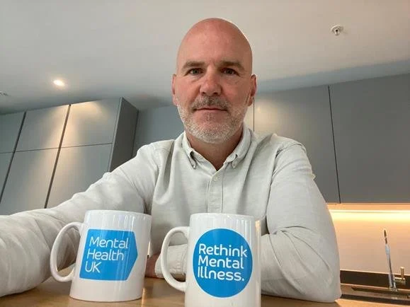 A photo of Brian Dow wearing a white shirt sitting in a kitchen with Mental Health UK and Rethink Mental Illness mugs in front of him.