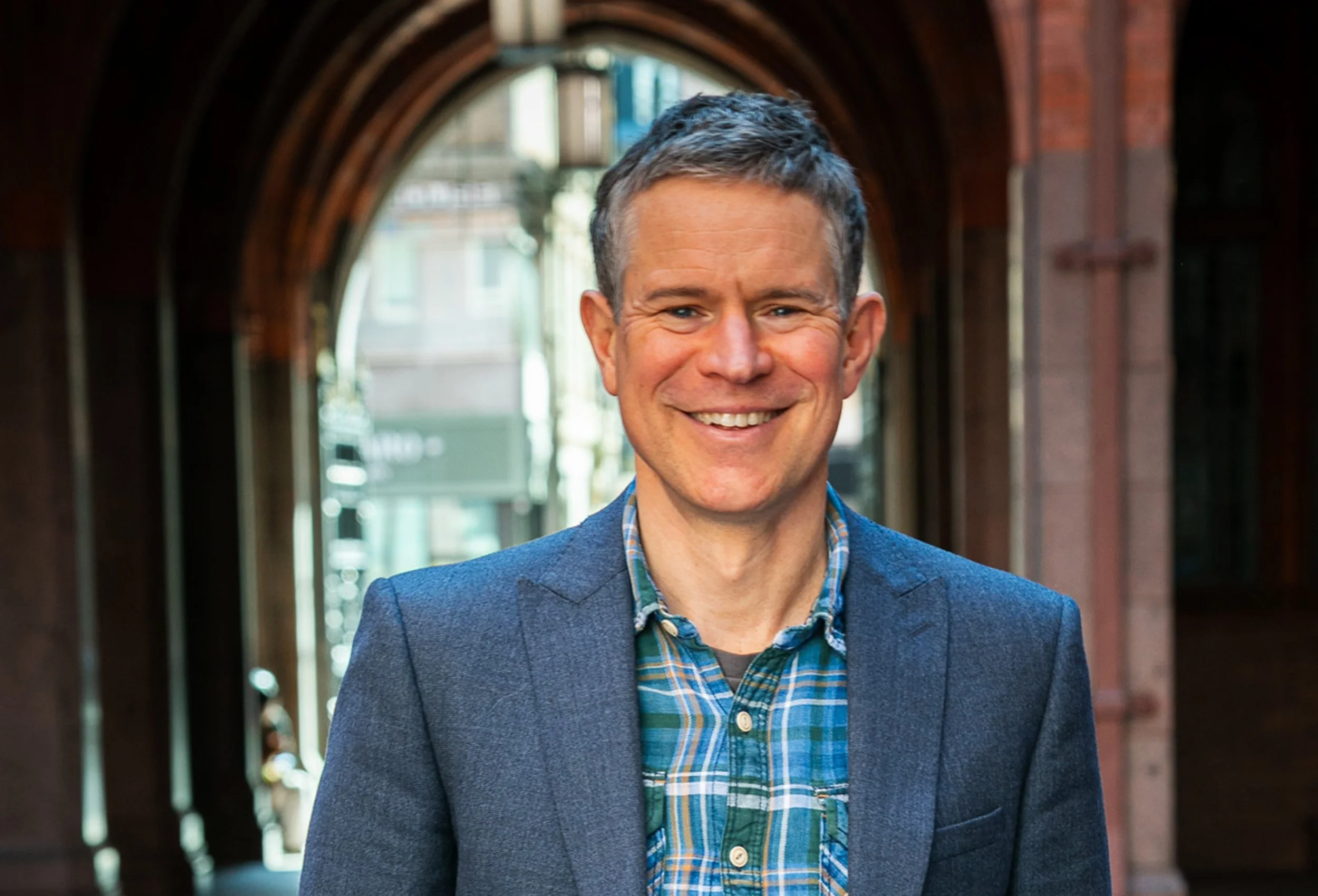 Portrait of Dr Mark Williamson smiling in a sunlit archway, wearing a blazer and checked shirt