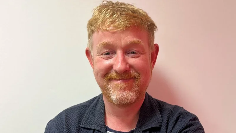 A photo of Chris Sherwood, a man with blonde hair and a beard, wearing a blue jacket and standing in front of a white wall
